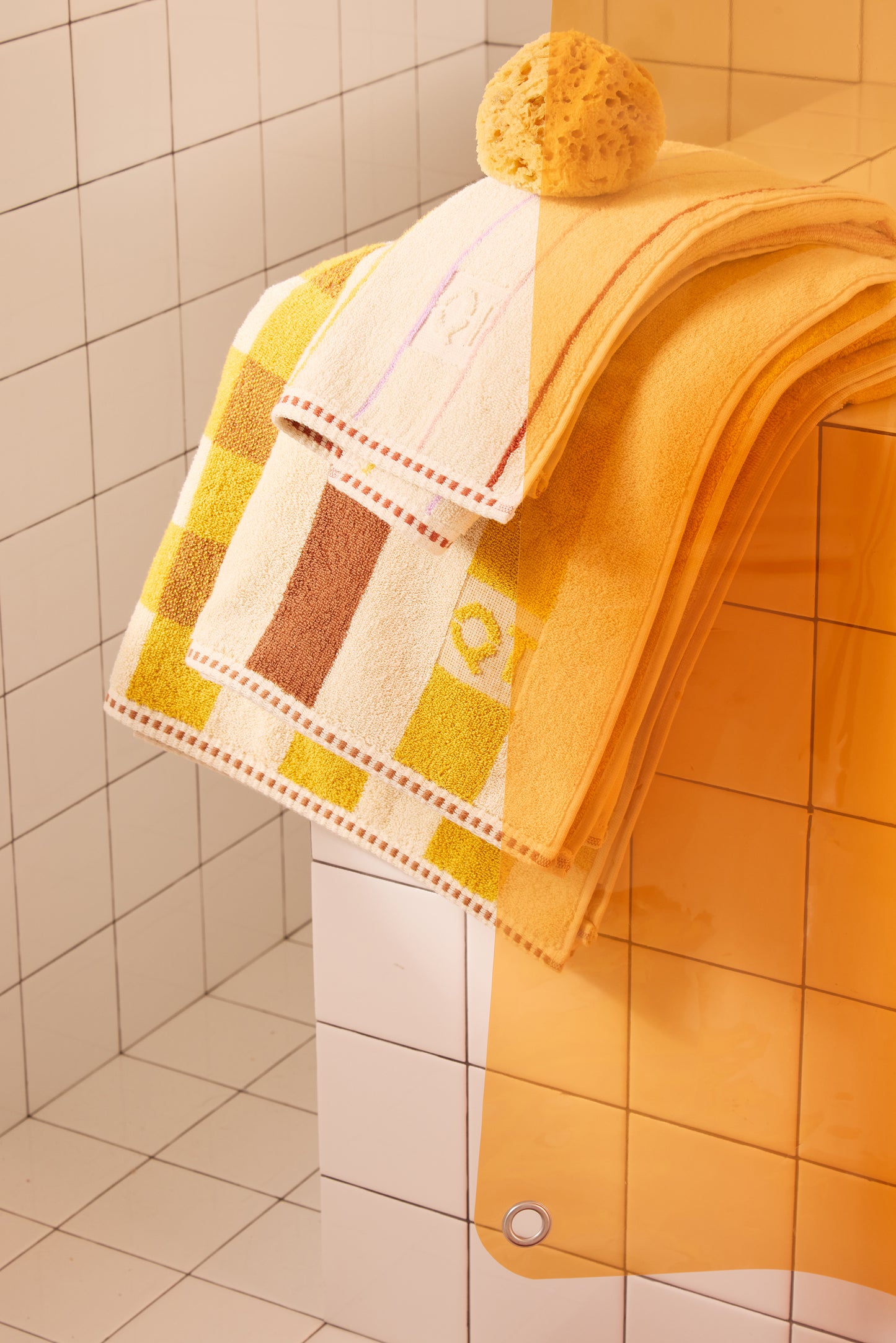 Yellow towels and orange clear curtain with a sponge in a tiled bathroom.