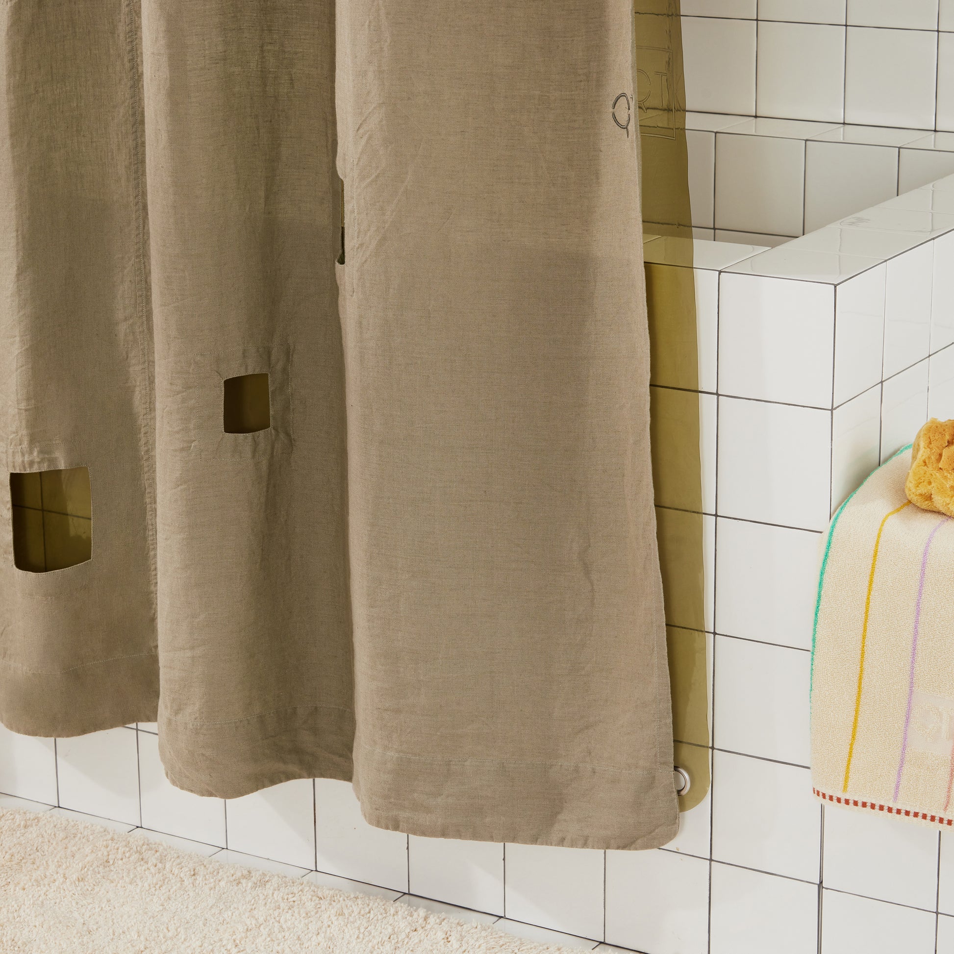 Bottom right corner of ronchamp linen curtain in olive against white tiled bathtub.