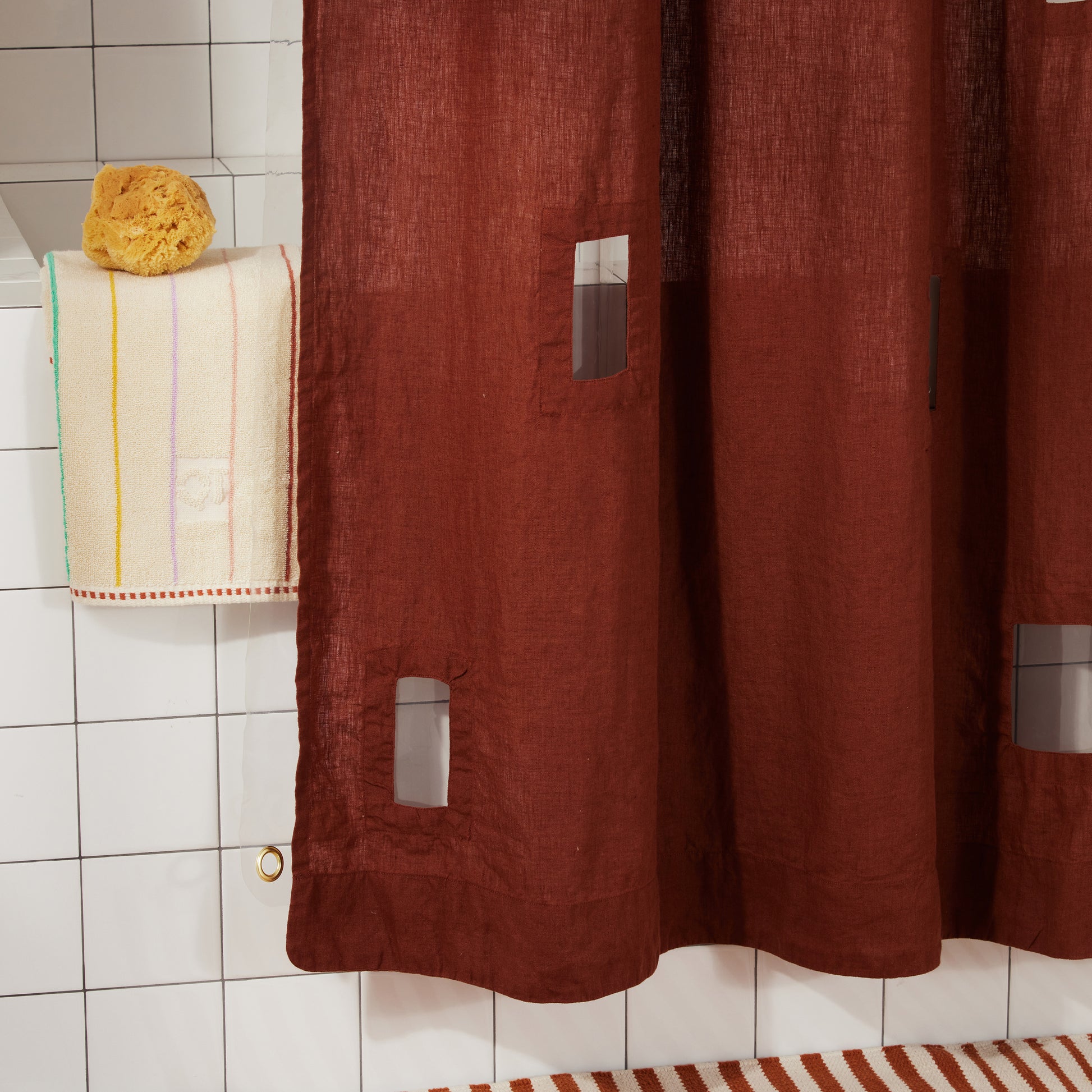 Close-up of bottom corner of burgundy linen shower curtain with cutout window detail against white tiled shower.