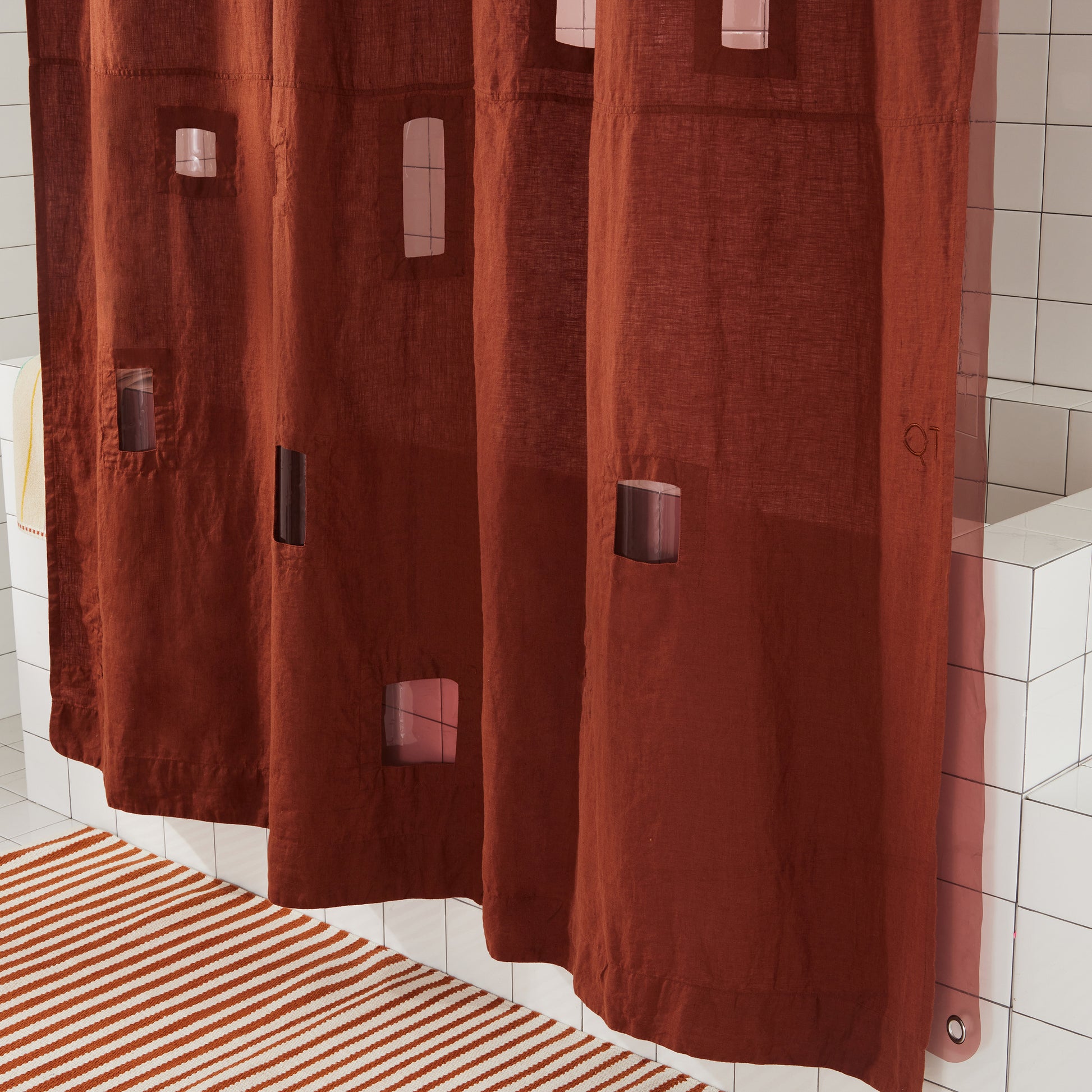 Bottom portion of burgundy linen shower curtain with assorted window cut outs, hanging above burgundy and white striped bath mat.