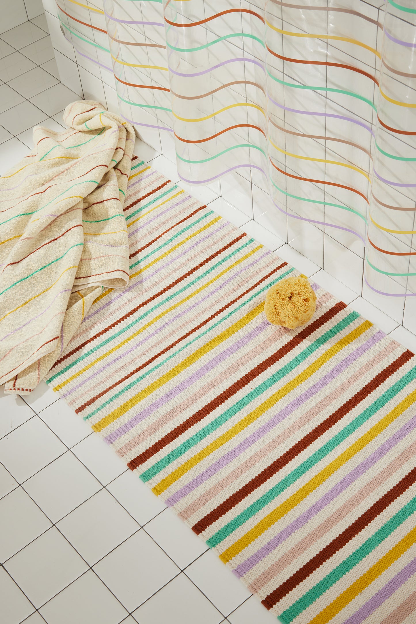 Multi colored striped prism rug and prism towels are styled laying flat on a bathroom floor with the prism sun shower curtain.