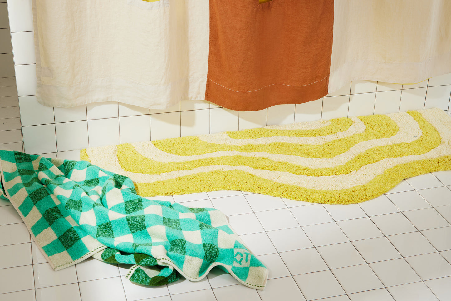 Yellow and cream wavy bath runner laying flat on white bathroom tile with clay and cream linen curtain and green checker towel on floor.