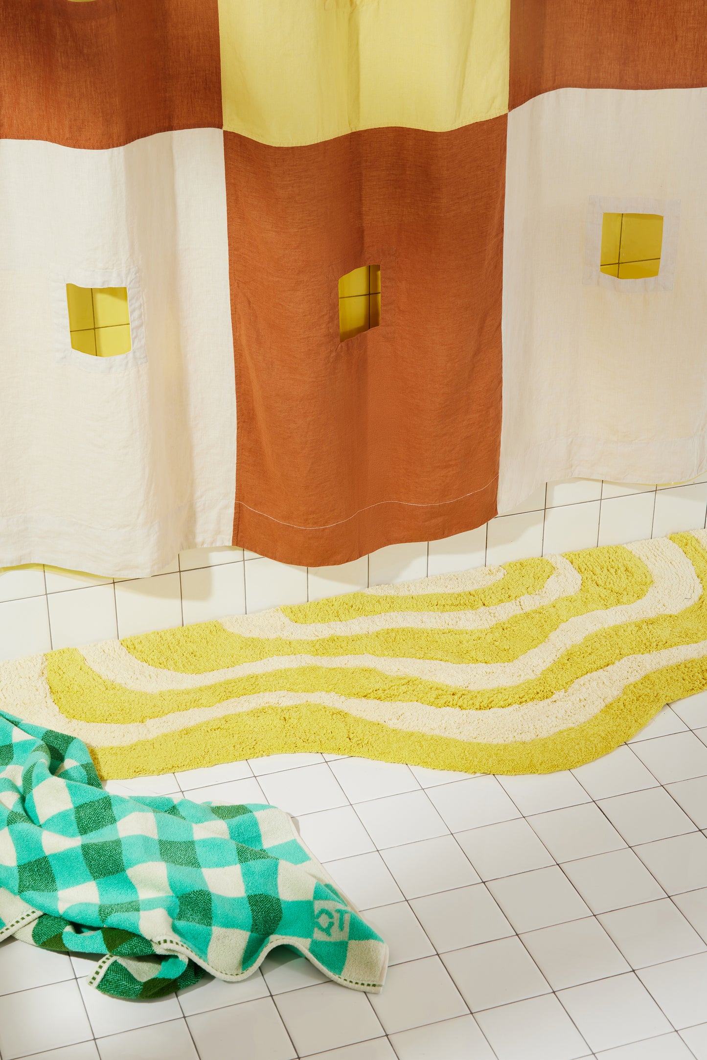 Yellow and cream wavy bath runner laying flat on white bathroom tile with clay and cream linen curtain and green checker towel on floor.