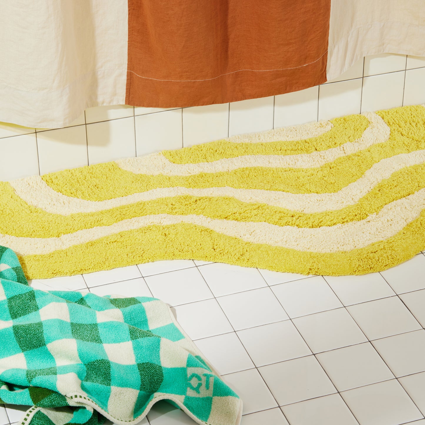 Yellow and cream wavy bath runner laying flat on white bathroom tile with clay and cream linen curtain and green checker towel on floor.