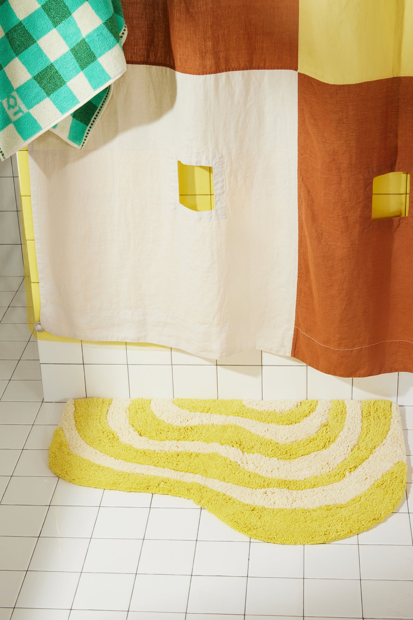 Yellow and cream wavy bath rug laying flat on white bathroom tile with clay and cream linen curtain and yellow sun shower layered behind curtain.