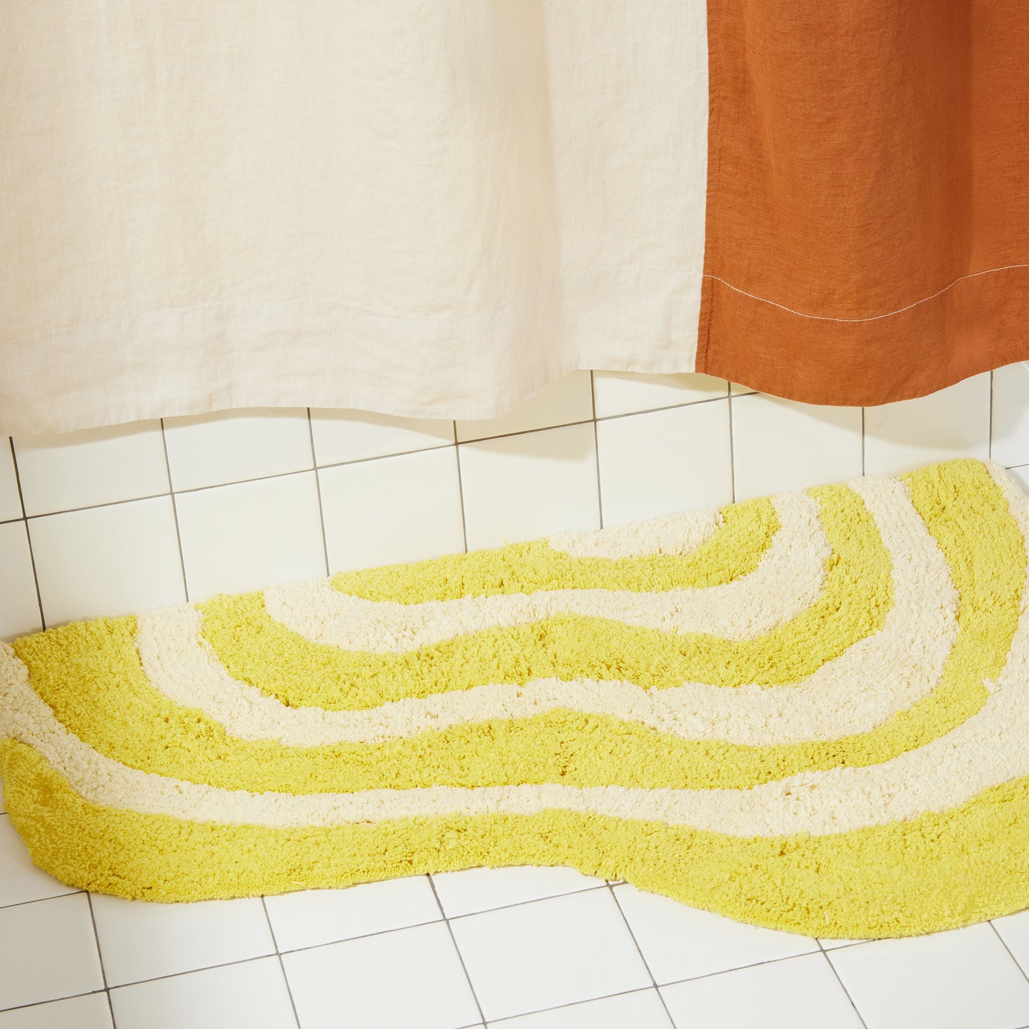Yellow and cream wavy bath rug laying flat on white bathroom tile with clay and cream linen curtain.