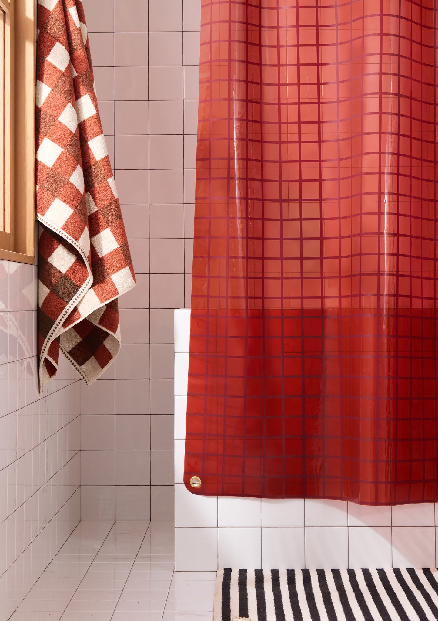 Bottom left corner of sun shower curtain in brick red styled with brown towel and black and white striped bath mat.