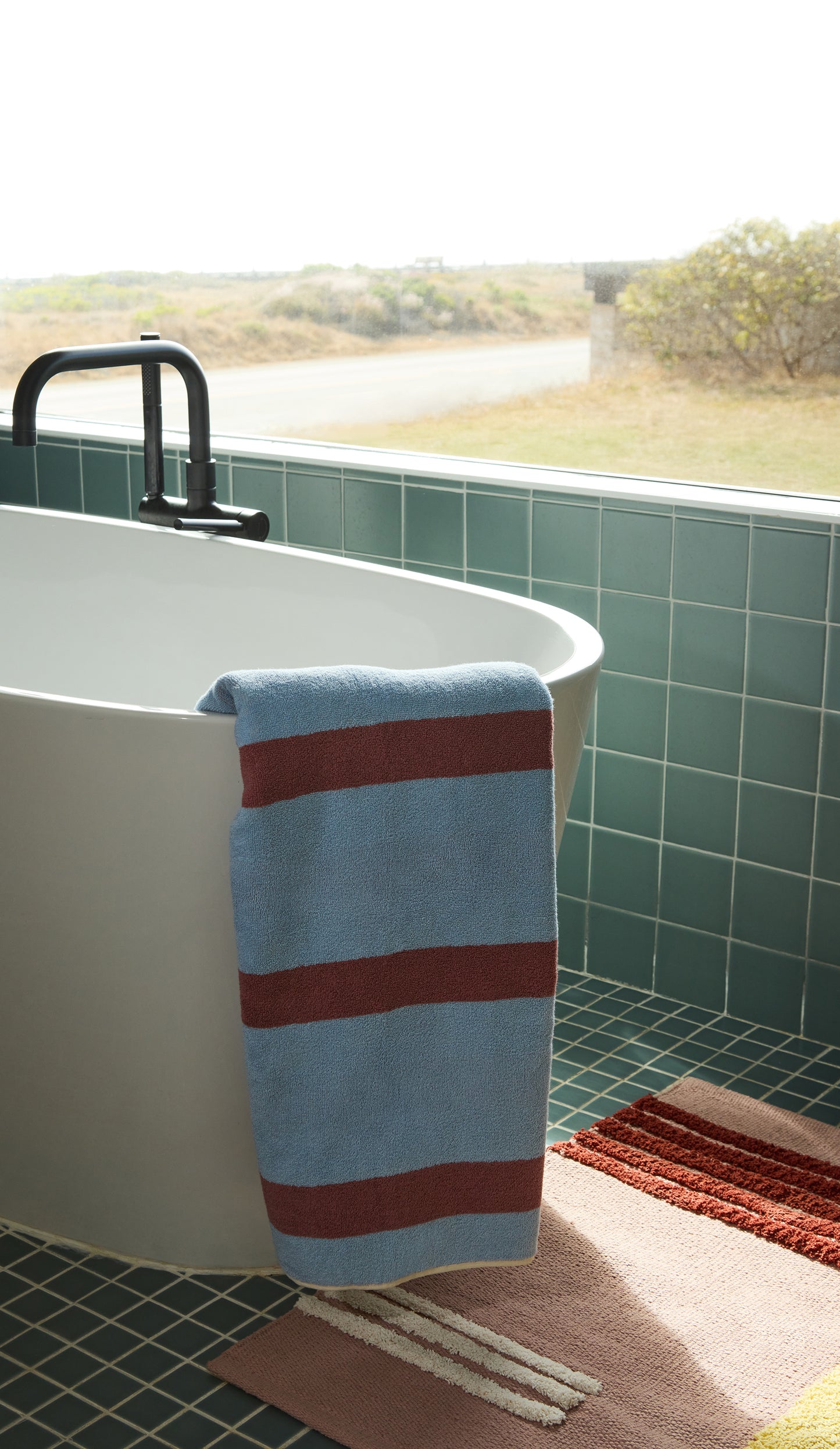 Striped blue and rose towel hanging over a white bathtub with blue tiled walls and a view of a road.