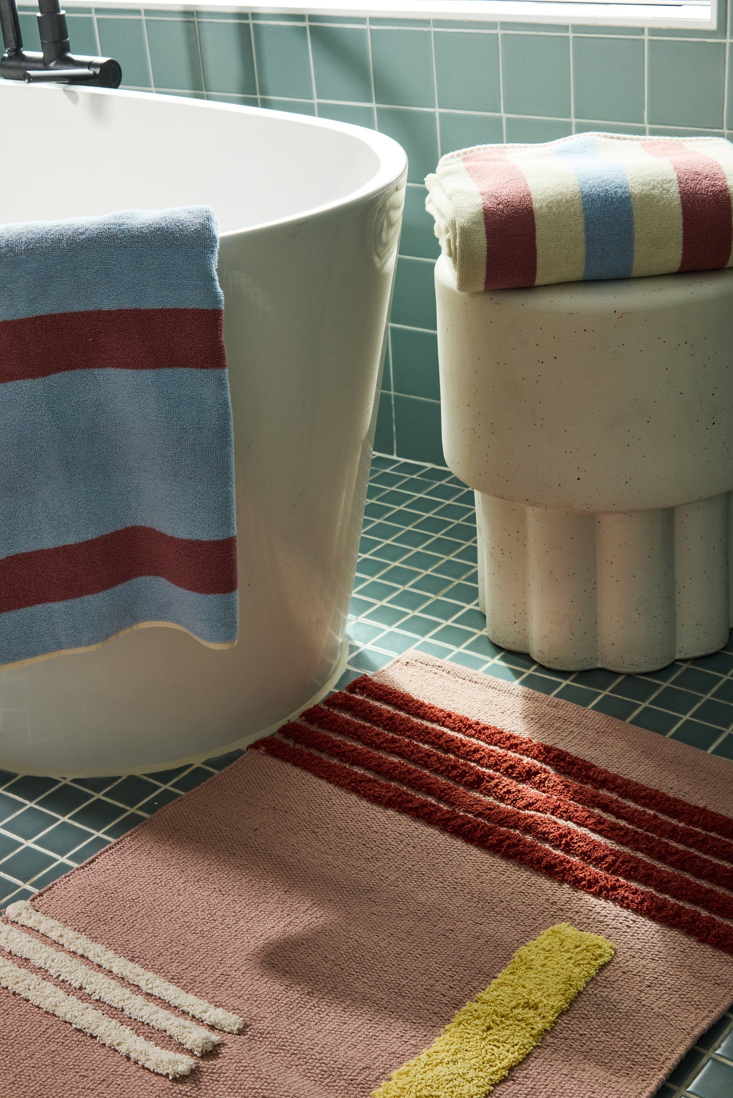 Bathroom scene with a bathtub, striped towel, and bath mat on a tiled floor.