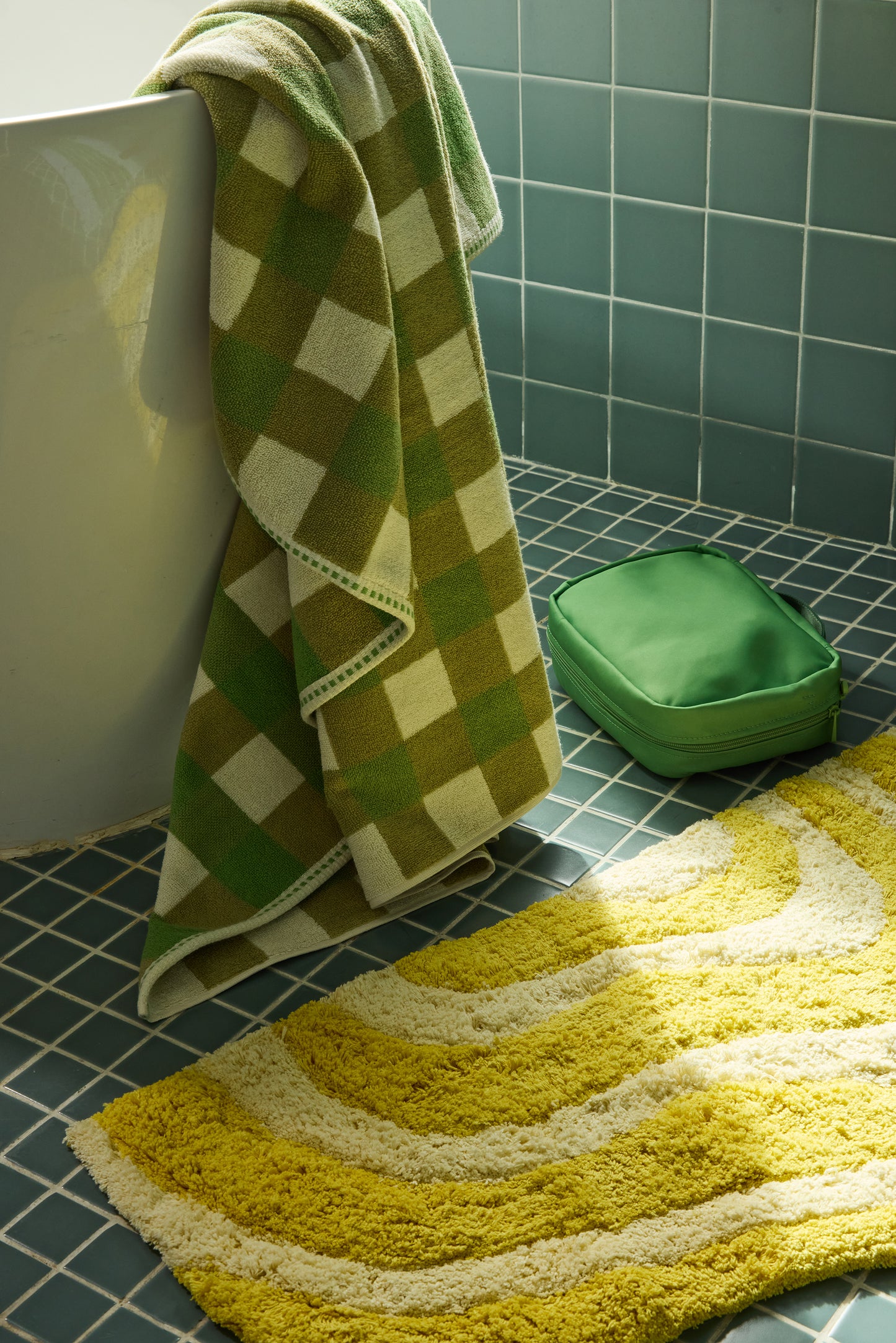 Green checkered towel draped over a white tub with a green bag and yellow bath mat on a tiled floor.
