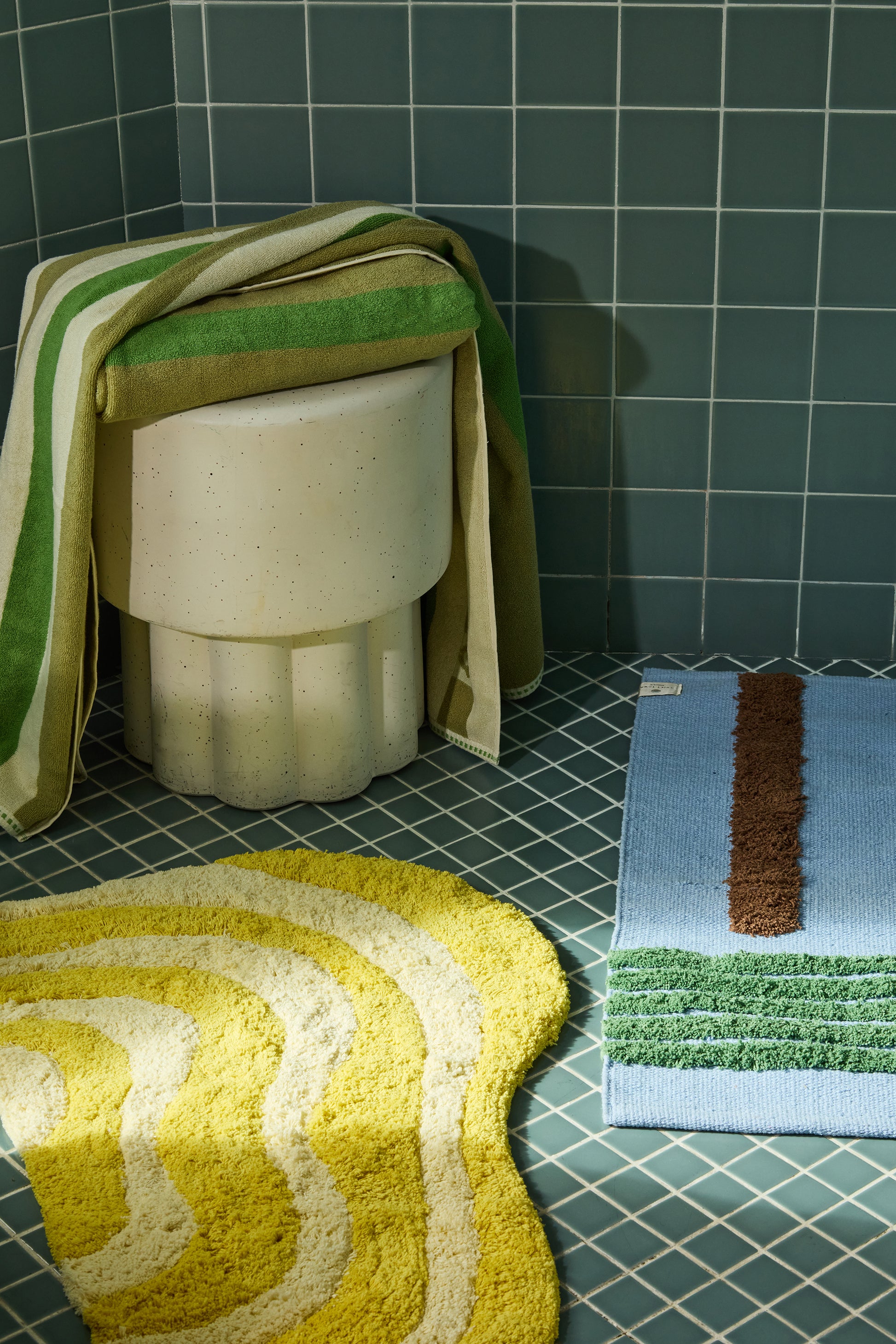 green and cream striped towels folded over stool surrounded by Bath mats in various colors on a tiled bathroom floor.