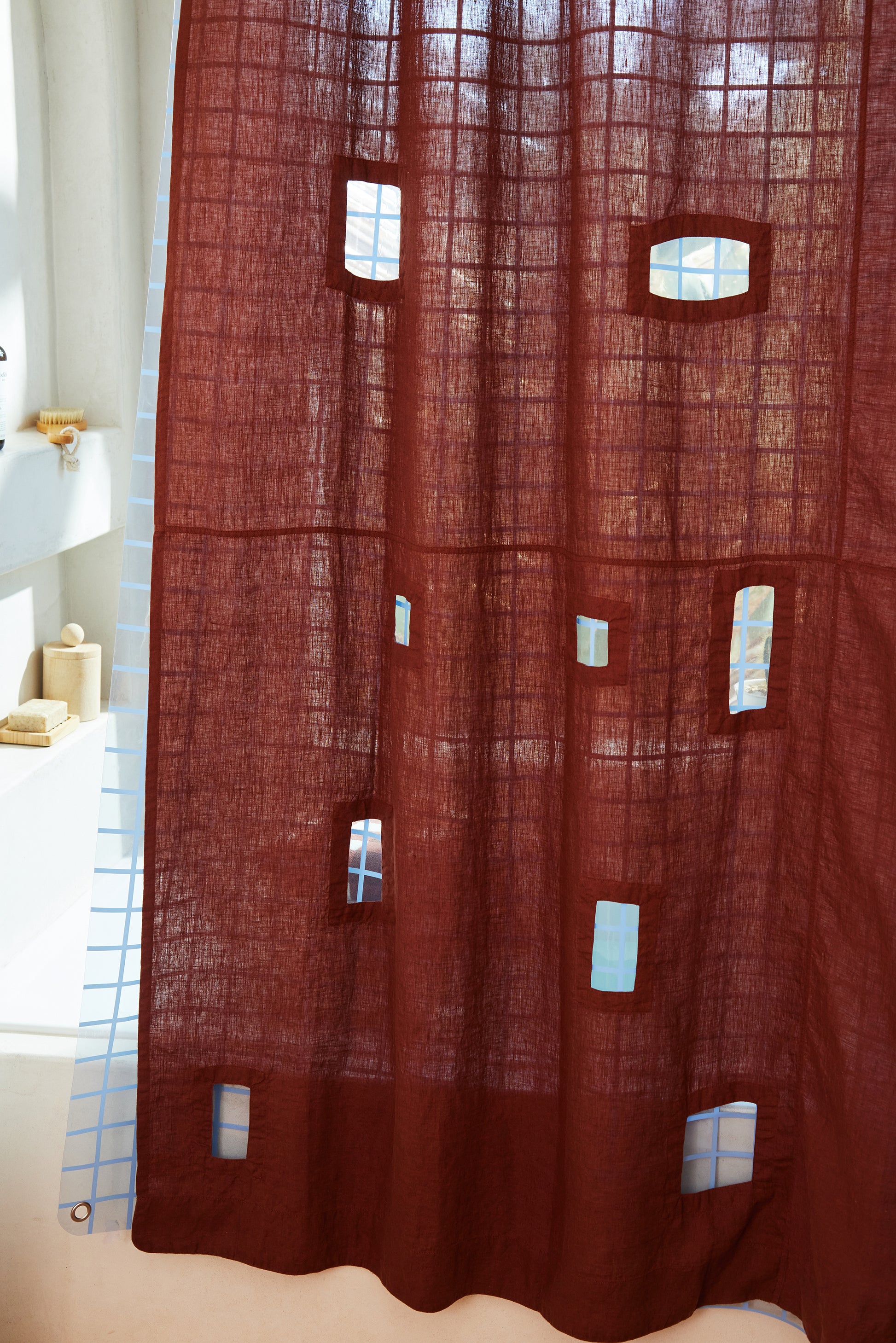 Burgundy linen shower curtain with rectangular window cut outs hanging over 2x2 blue sun shower curtain in a light filled bathroom to detail visible layering.