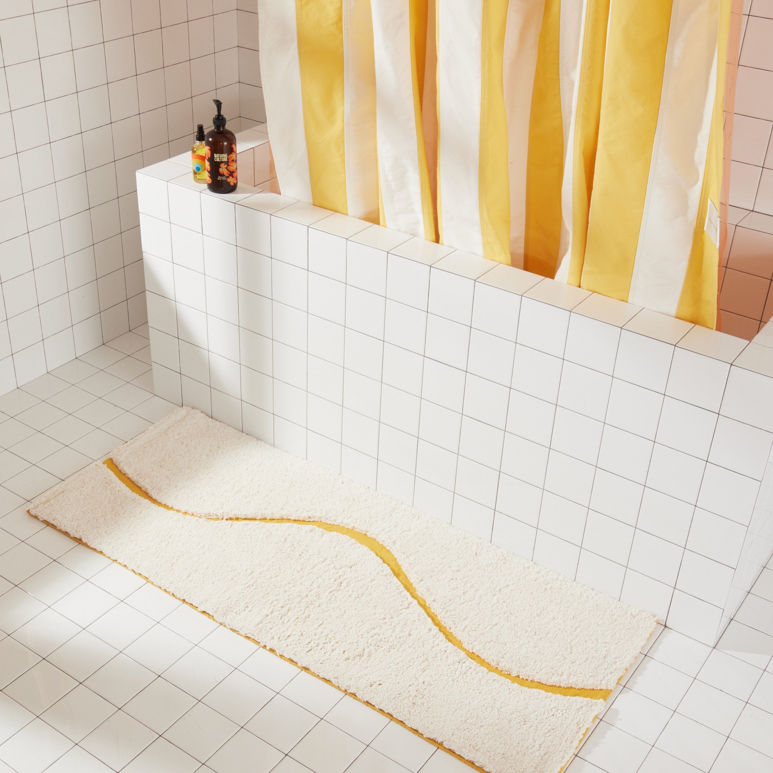 Full view of white bath mat in 56"x22" size with yellow curvy path stripe contrast styled in a white tiled bathroom.