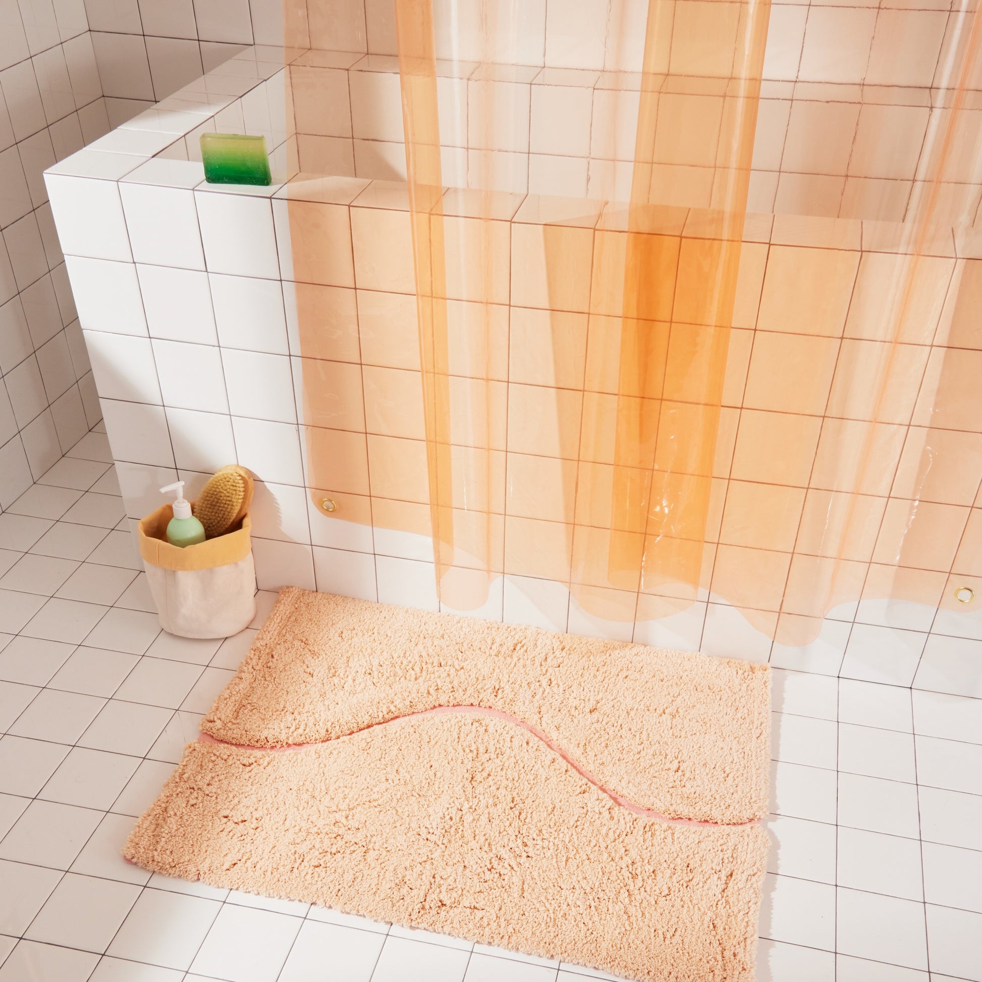 Peach bath mat in 21"x31" size with pink curvy path stripe contrast styled next to white tiled bathtub and below a peach sun shower curtain.