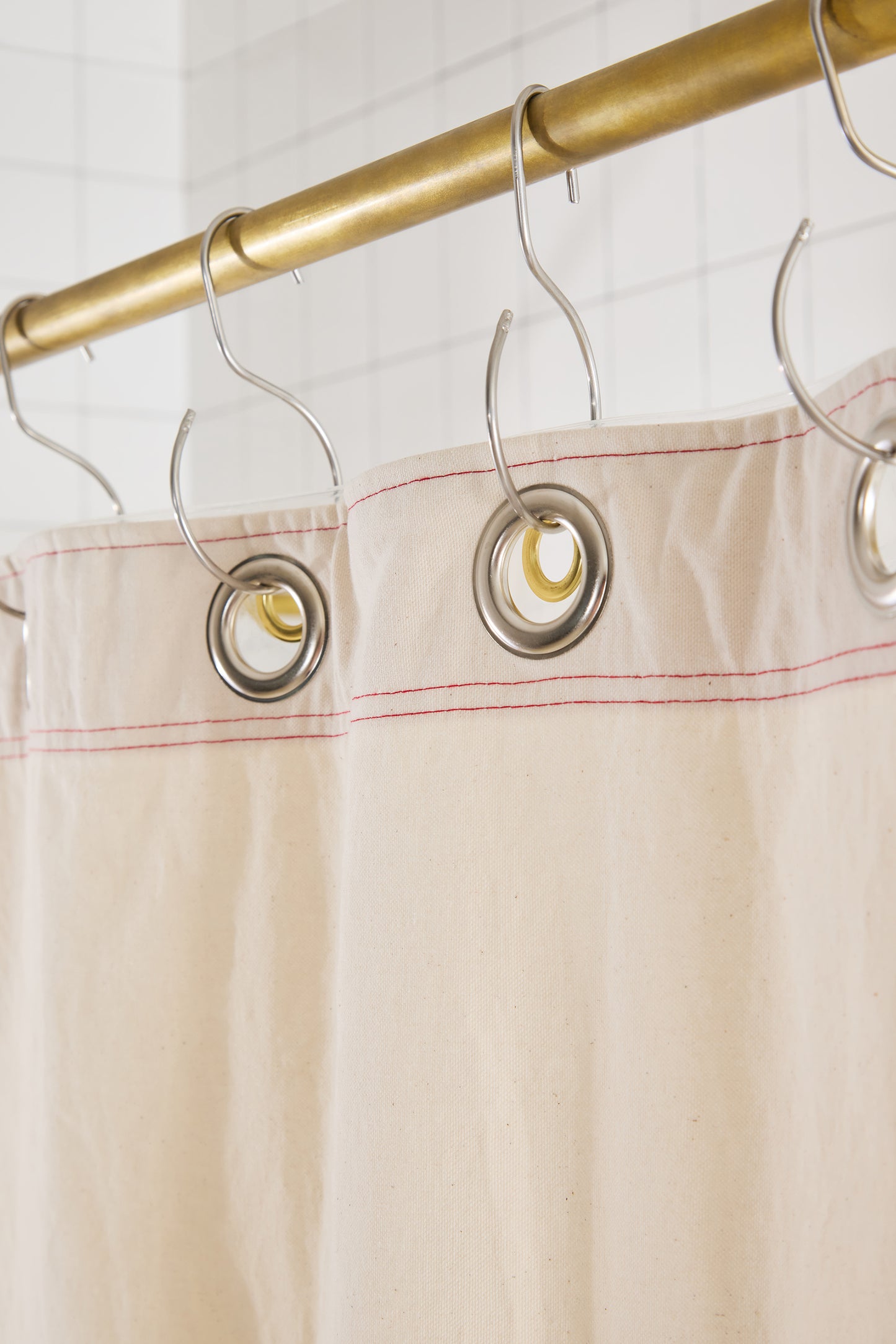 cream curtain with red stitching and large silver grommets hanging from silver hooks on a brass rod. 