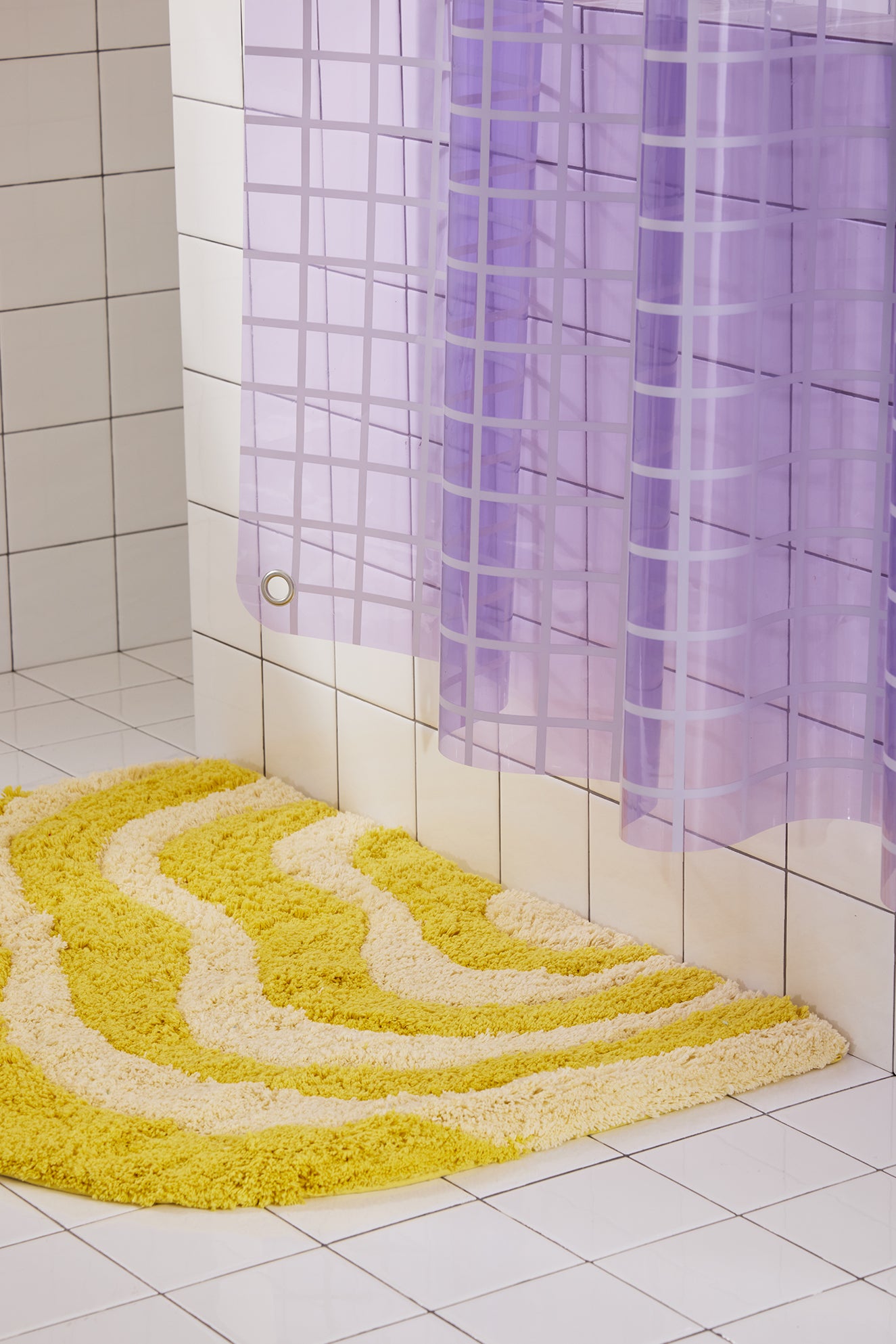 Yellow textured bath mat on a tiled bathroom floor with a lavender shower curtain.