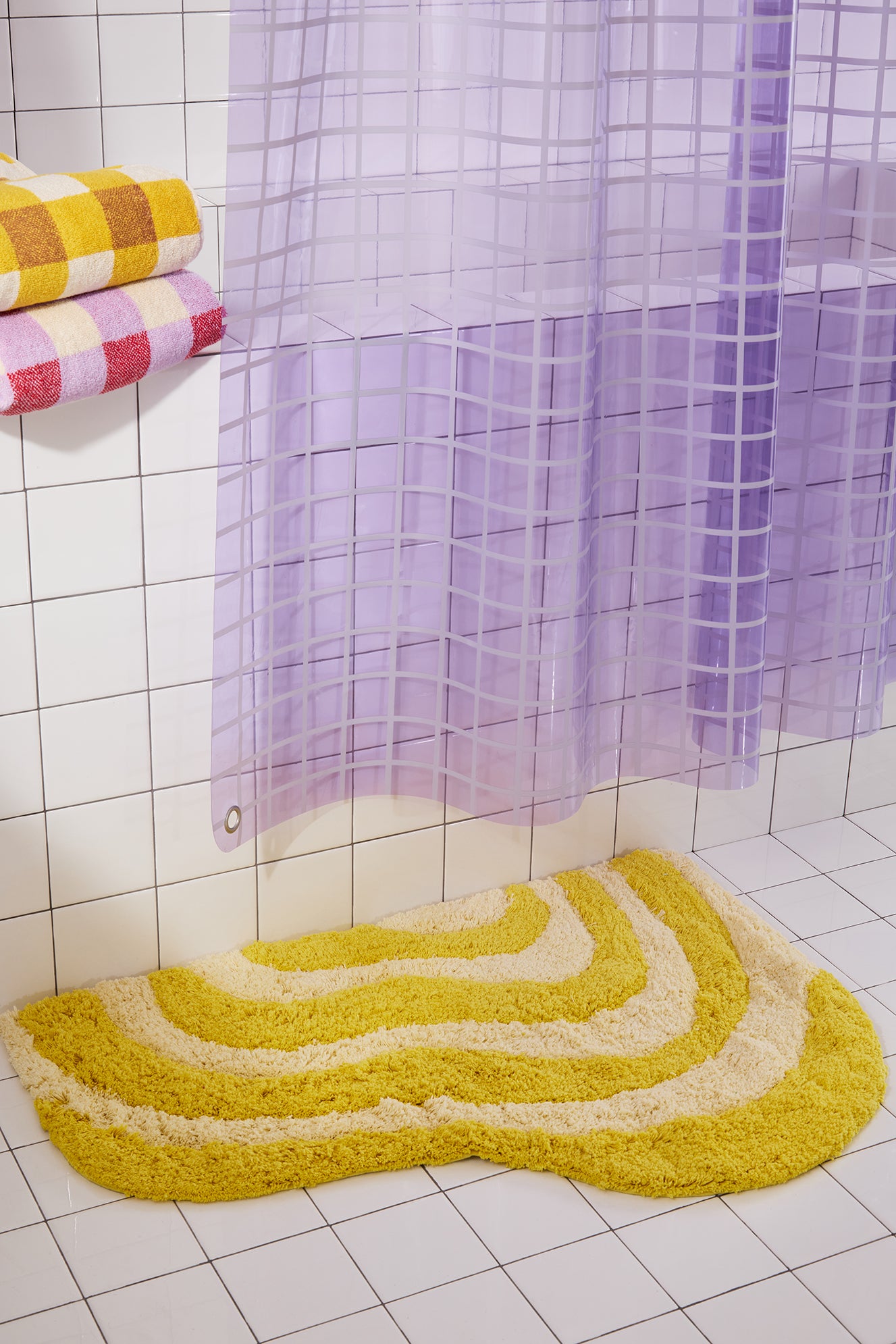 Bathroom with a yellow bath mat, purple shower curtain, and colorful towels on a tiled wall.