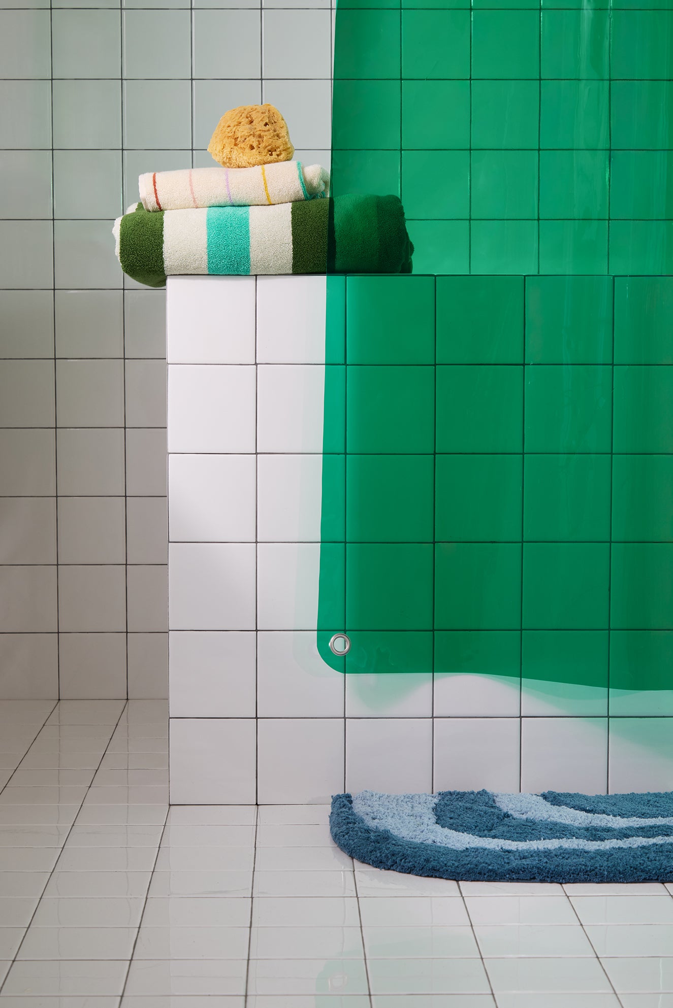 Green shower curtain with a bluerug and towels in a tiled bathroom.
