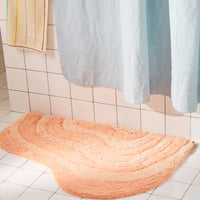 Peach curvy bath mat on a tiled bathroom floor with a shower curtain in the background.