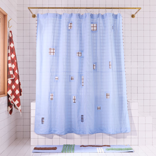 Blue curtain with cut outs and check liner styled behind it in a white tiled bathroom. 