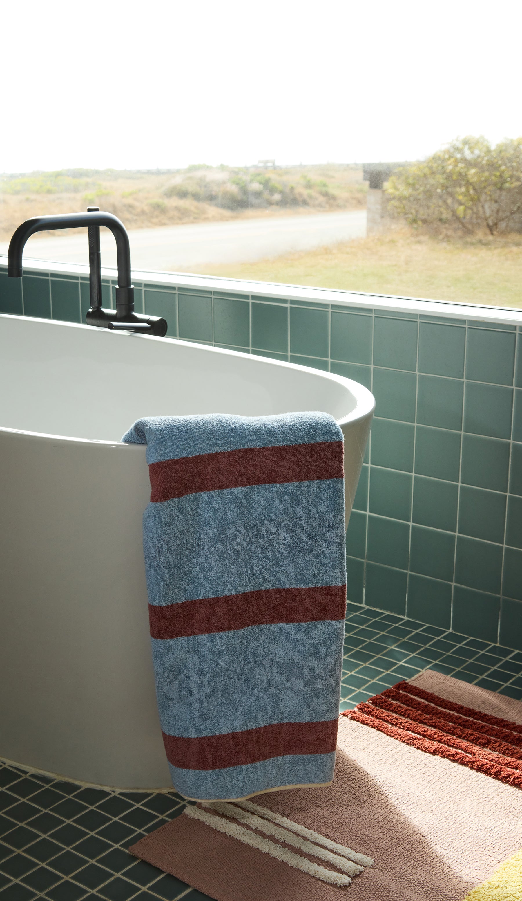 Striped blue and rose towel hanging over a white bathtub with blue tiled walls and a view of a road.