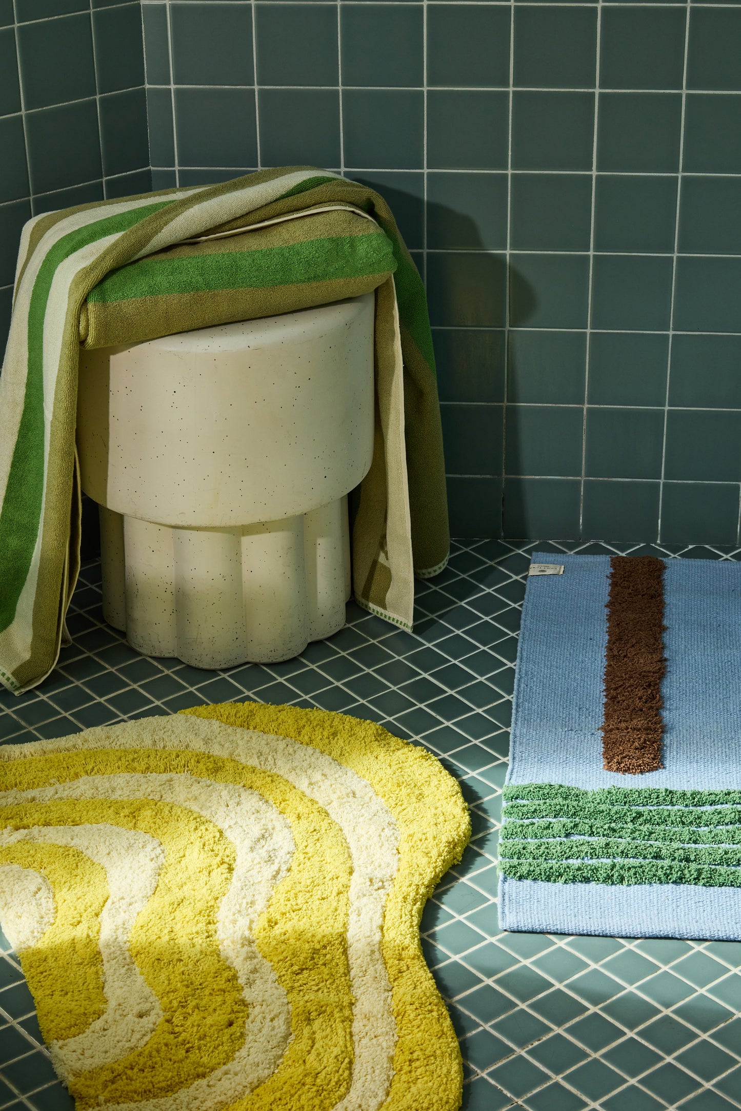 green and cream striped towels folded over stool surrounded by Bath mats in various colors on a tiled bathroom floor.
