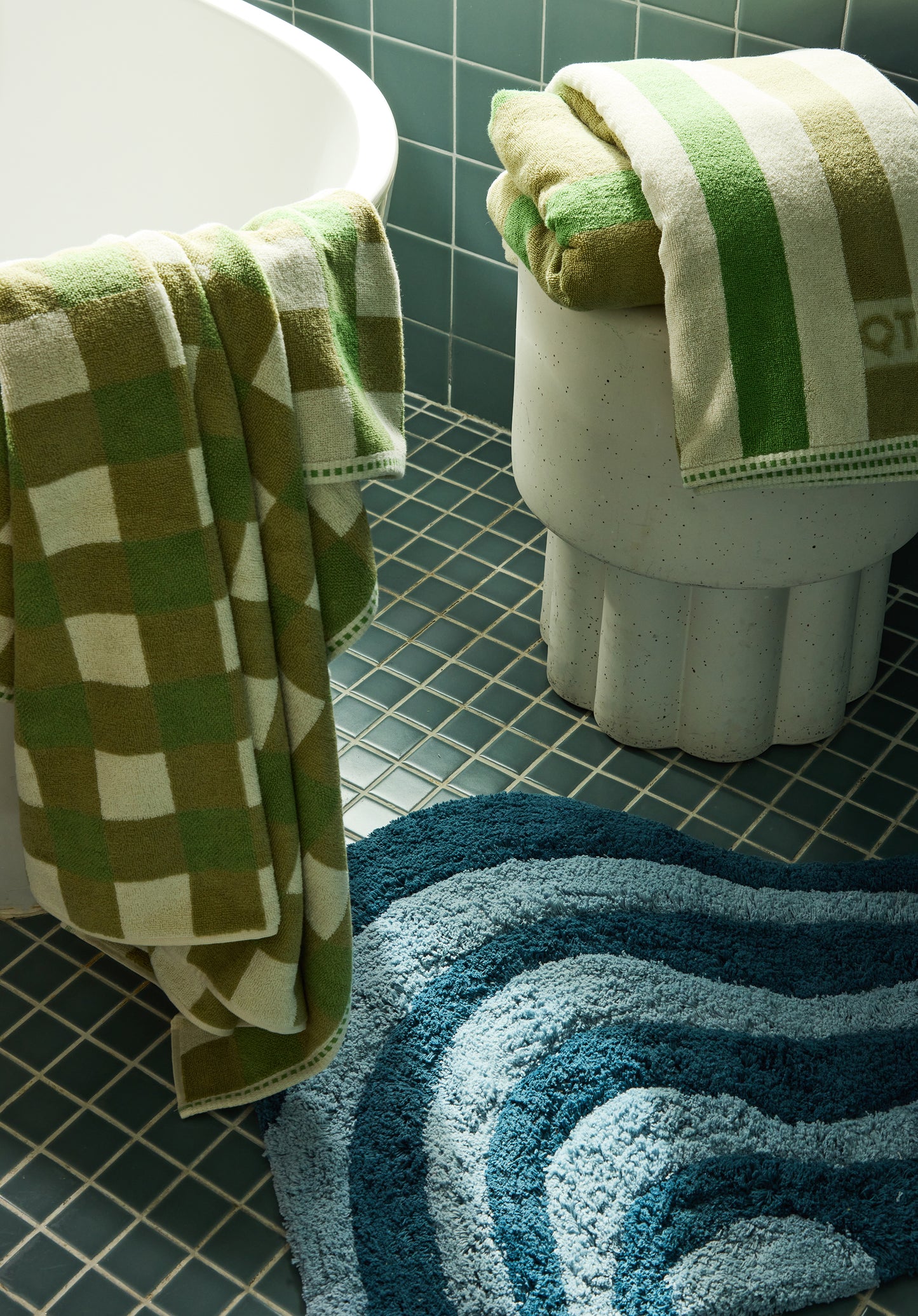 Bathroom with green and white checkered towel, striped towel, and blue textured rug on a tiled floor.