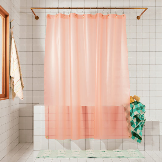 A peach-pink translucent shower curtain hanging from a brass rod in a tiled bathroom, with a green plaid towel draped over the tub edge and a mint bath mat on the floor.