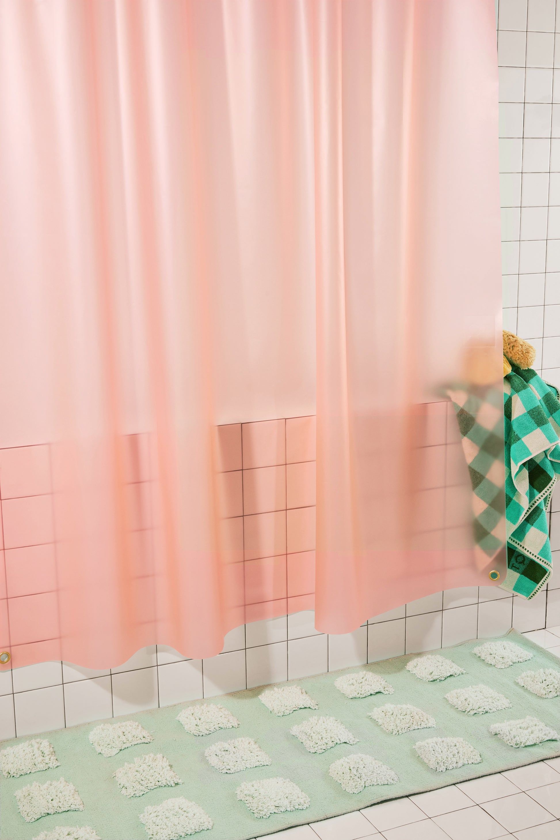 A pastel pink translucent shower curtain hanging in a tiled bathroom, with a mint green bath mat and a green-and-white checkered towel visible.