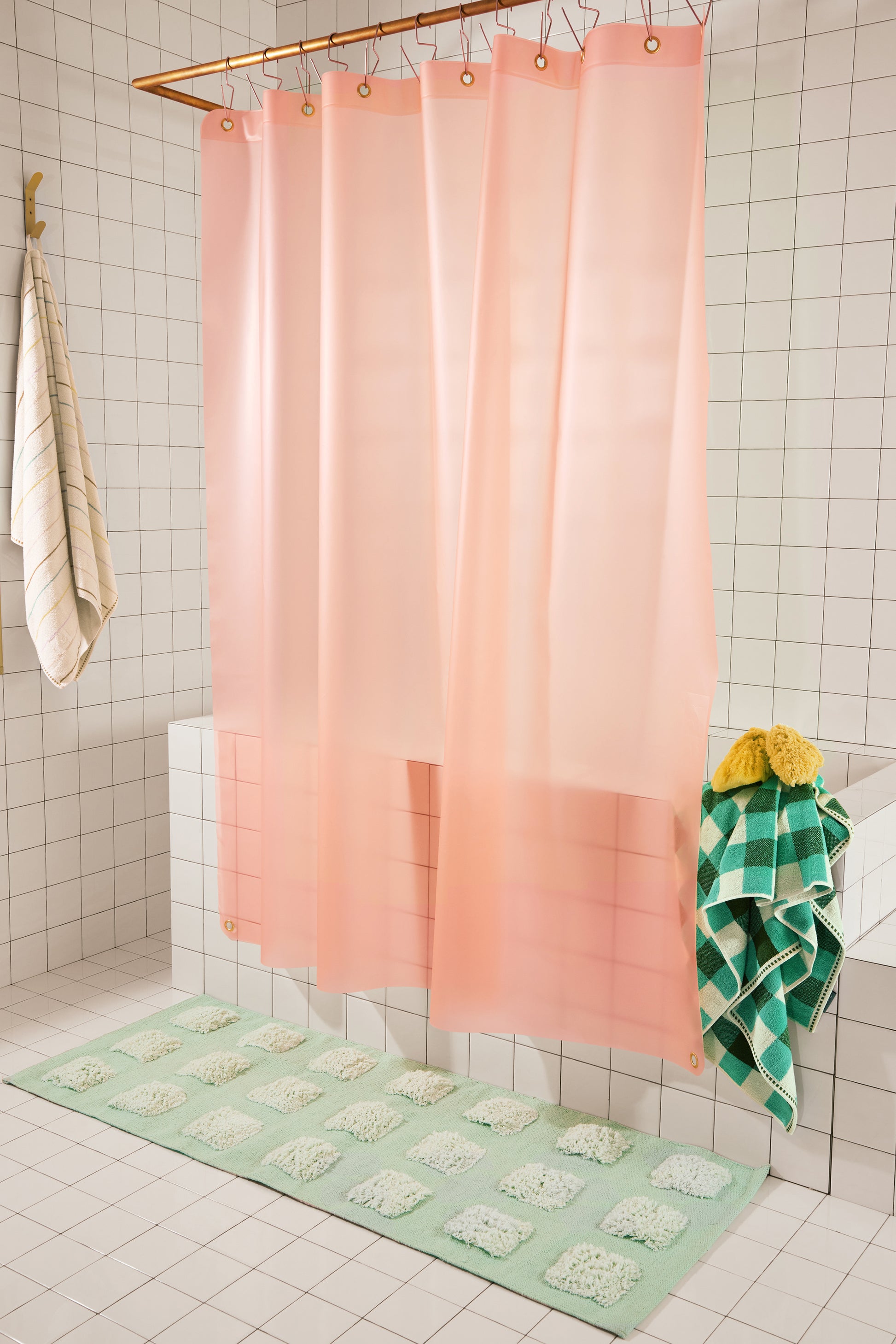 A peach-pink translucent shower curtain hanging from a gold rod in a tiled bathroom, with a mint green bath mat and a green-and-white checkered towel nearby.