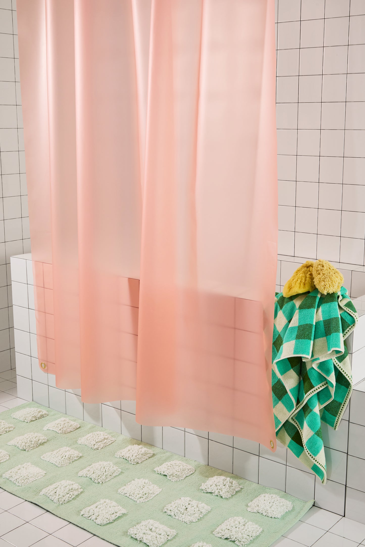 A peach-pink translucent shower curtain hanging in a tiled bathroom, with a green and white checkered towel draped over the side and a matching green bath mat on the floor.