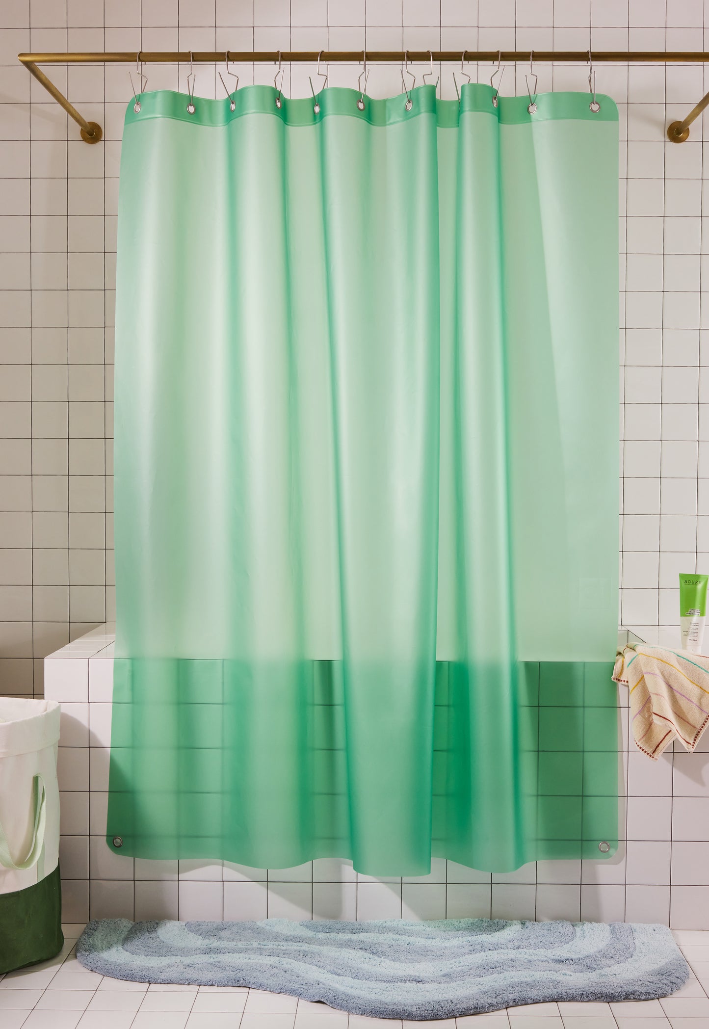 A mint green shower curtain hanging from a gold rod in a tiled bathroom, with a blue-gray bath mat on the floor and a towel on the right.