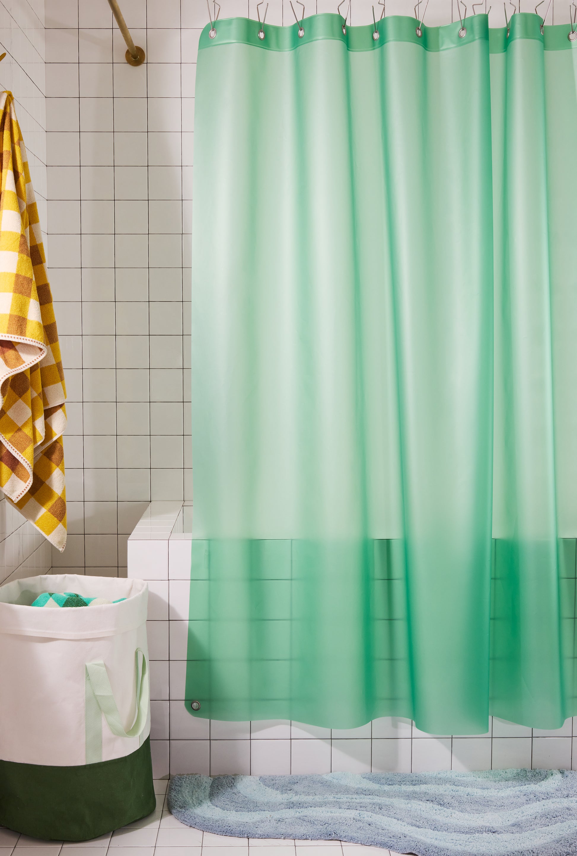 A mint green shower curtain hanging in a tiled bathroom, with a white tub, a yellow-and-white checkered towel, and a blue bath mat visible.