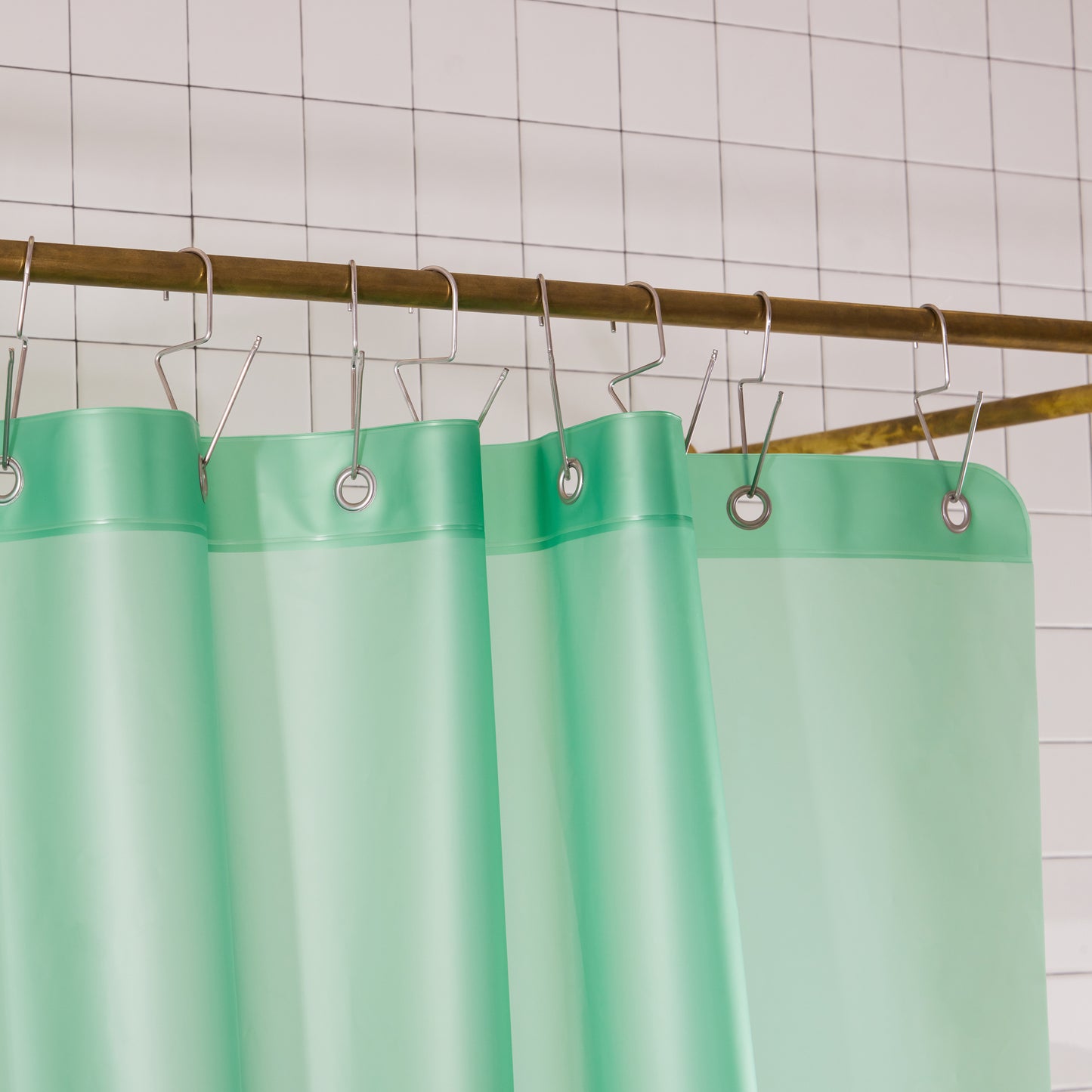 A mint green shower curtain hanging from silver metal hooks on a brass rod against a white tiled wall.