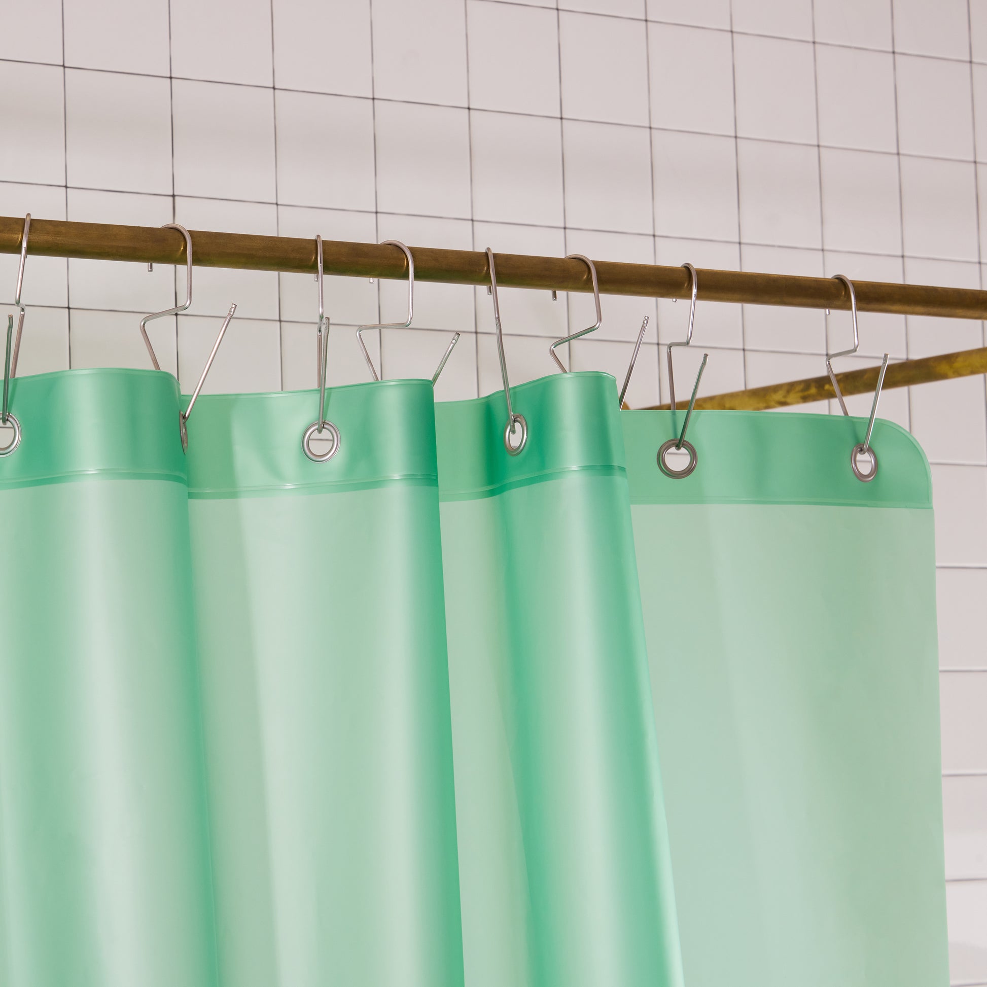 A mint green shower curtain hanging from silver metal hooks on a brass rod against a white tiled wall.