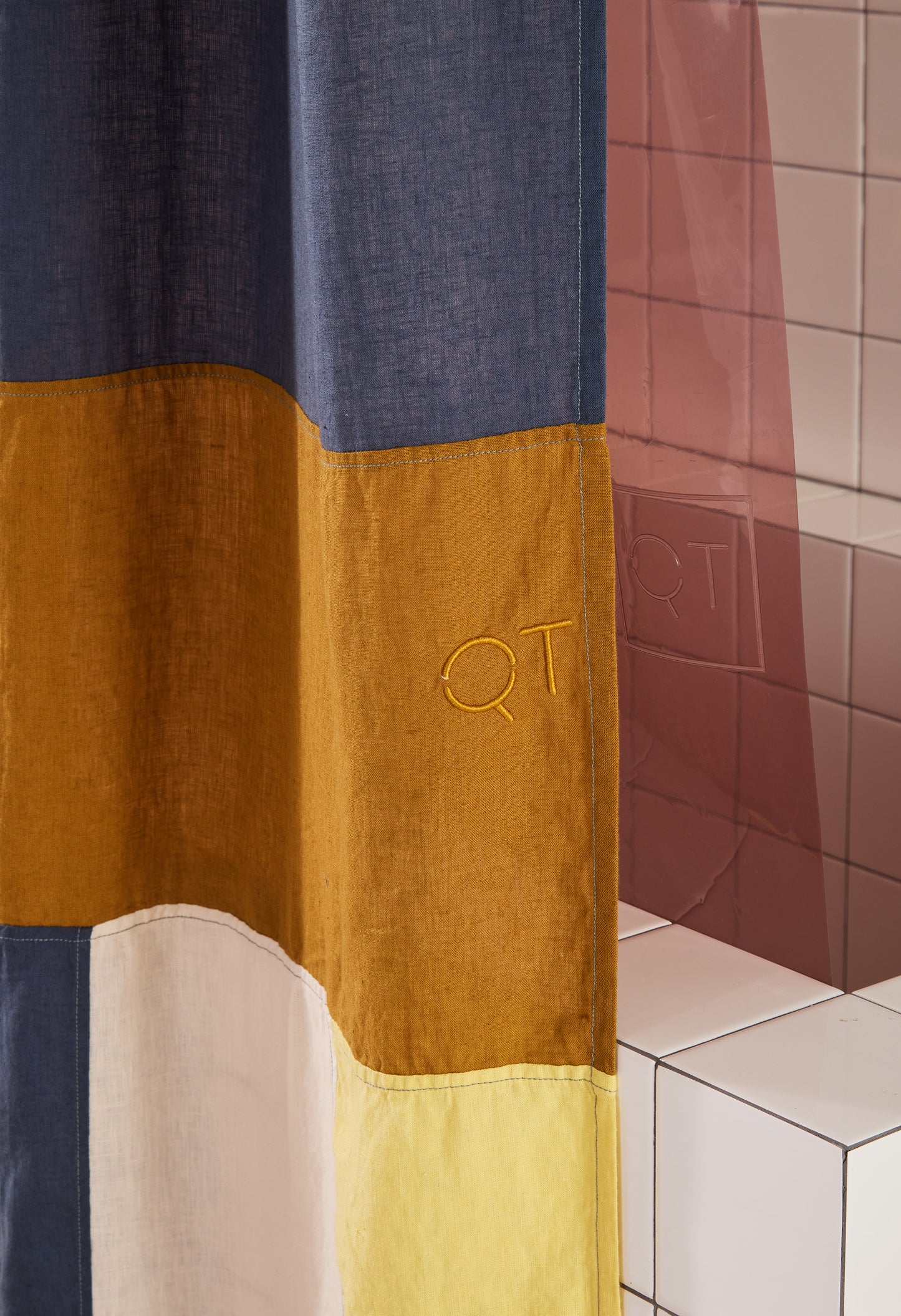 color-blocked linen curtain with QT embroidered and hanging in front of rose colored clear shower curtain.