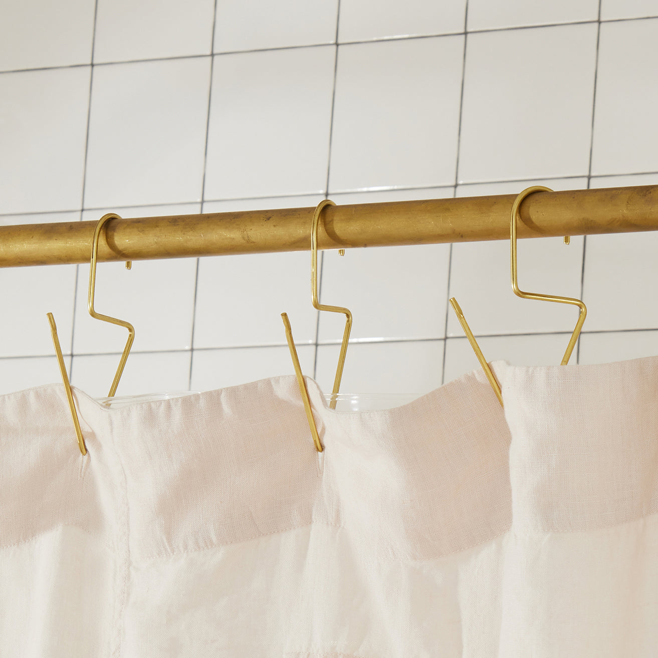 Gold hooks on a curtain rod with a white curtain against a tiled wall.