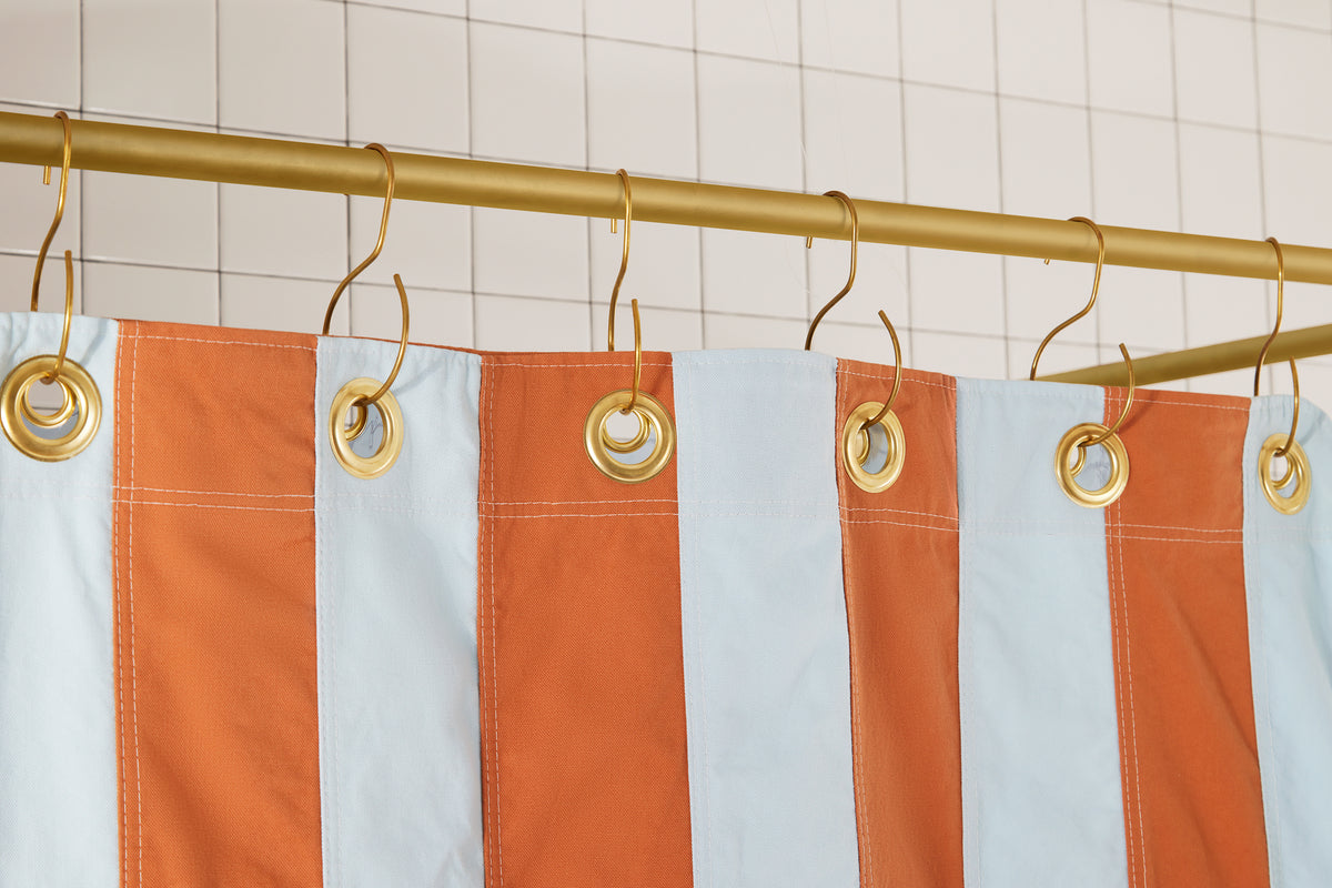 A striped shower curtain with alternating light blue and rust-orange vertical panels, hanging from gold metal grommets on a gold rod against a white tiled wall.