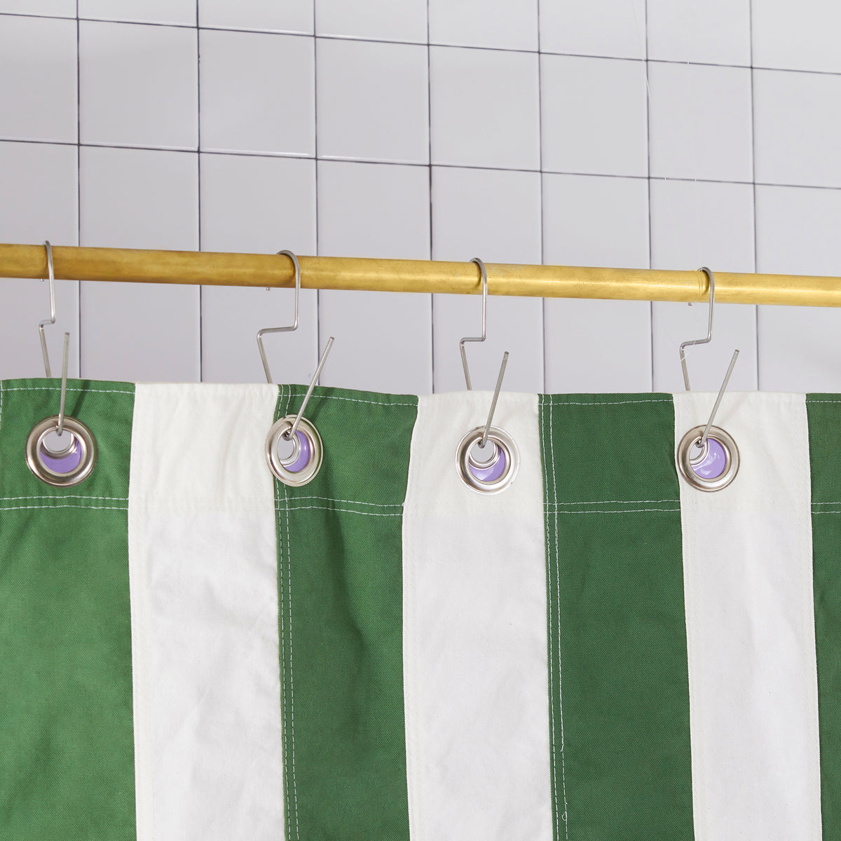Green and white striped shower curtain hanging from a gold metal rod with silver hooks against a white tiled wall.