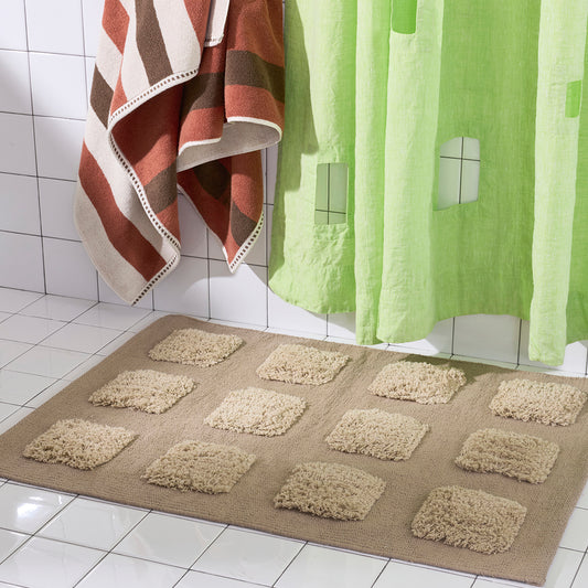 A rectangular bath mat with a grid of raised square cushions in a warm beige and  shag texture, shown on a white tiled bathroom floor with a striped towel and green shower curtain nearby.