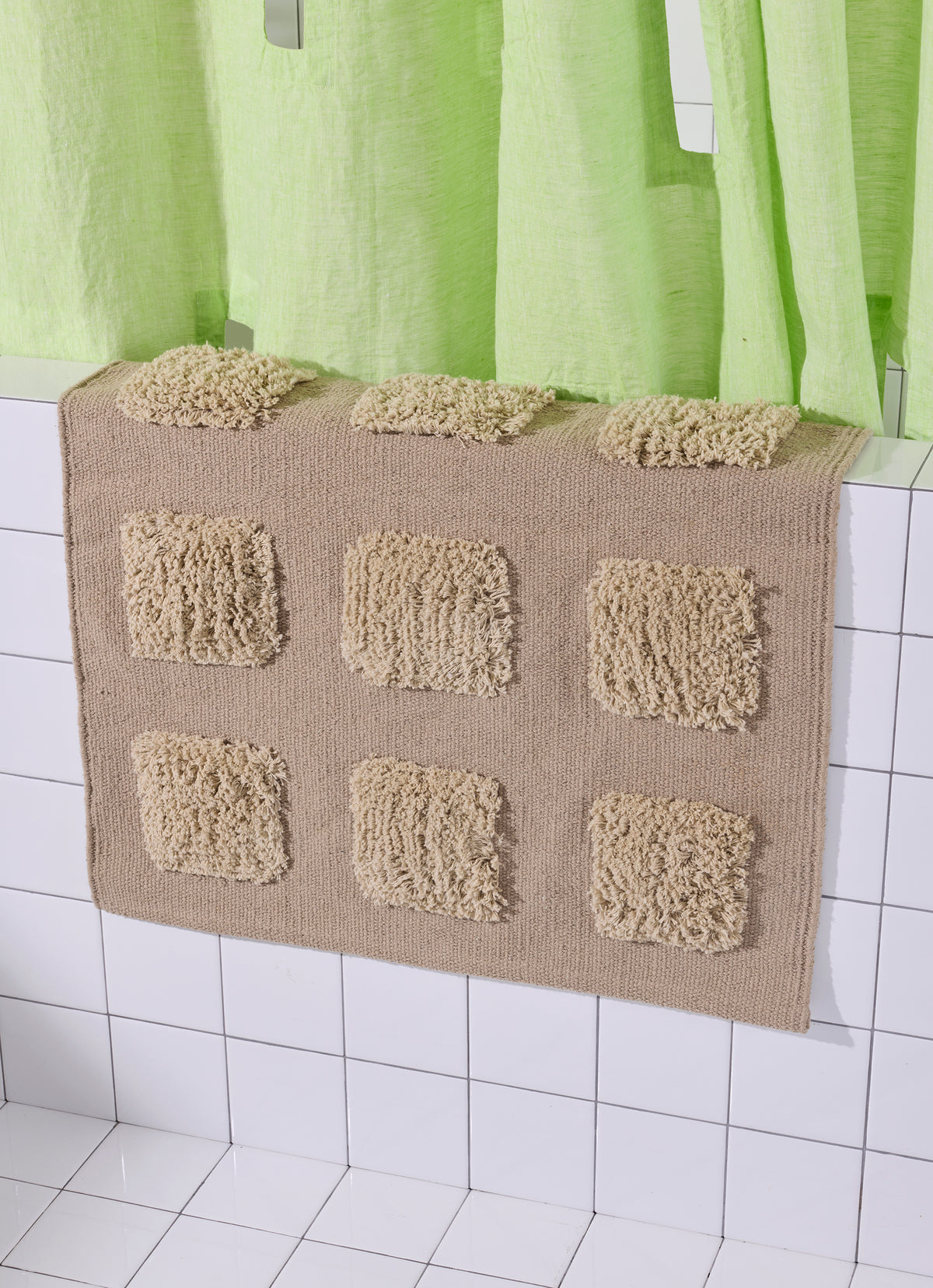 brown/taupe bath rug with raised tufted squares in natural tones, draped over a white tiled bathroom ledge beneath a lime green shower curtain.