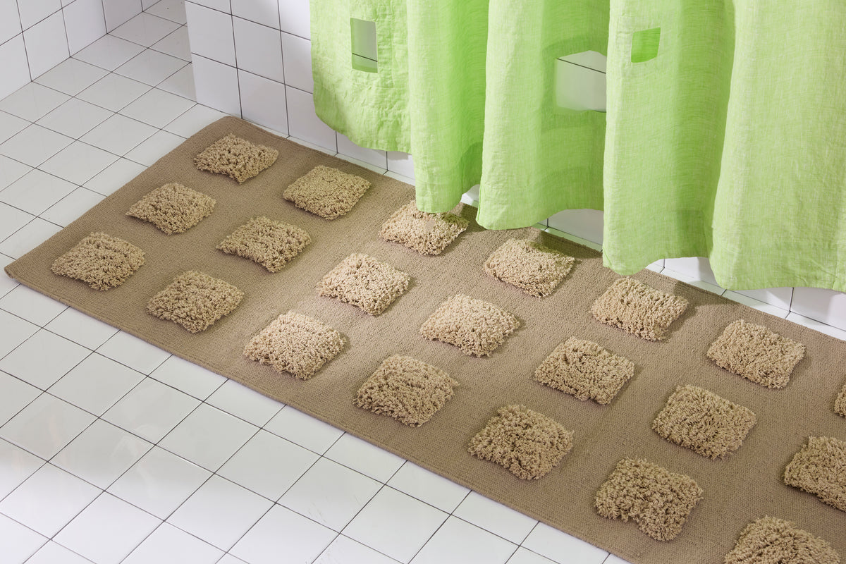 A brown/taupe, natural bath runner with an organic grid of raised tufted squares in warm neutral tones, shown in a bright white tiled bathroom with a lime green shower curtain.