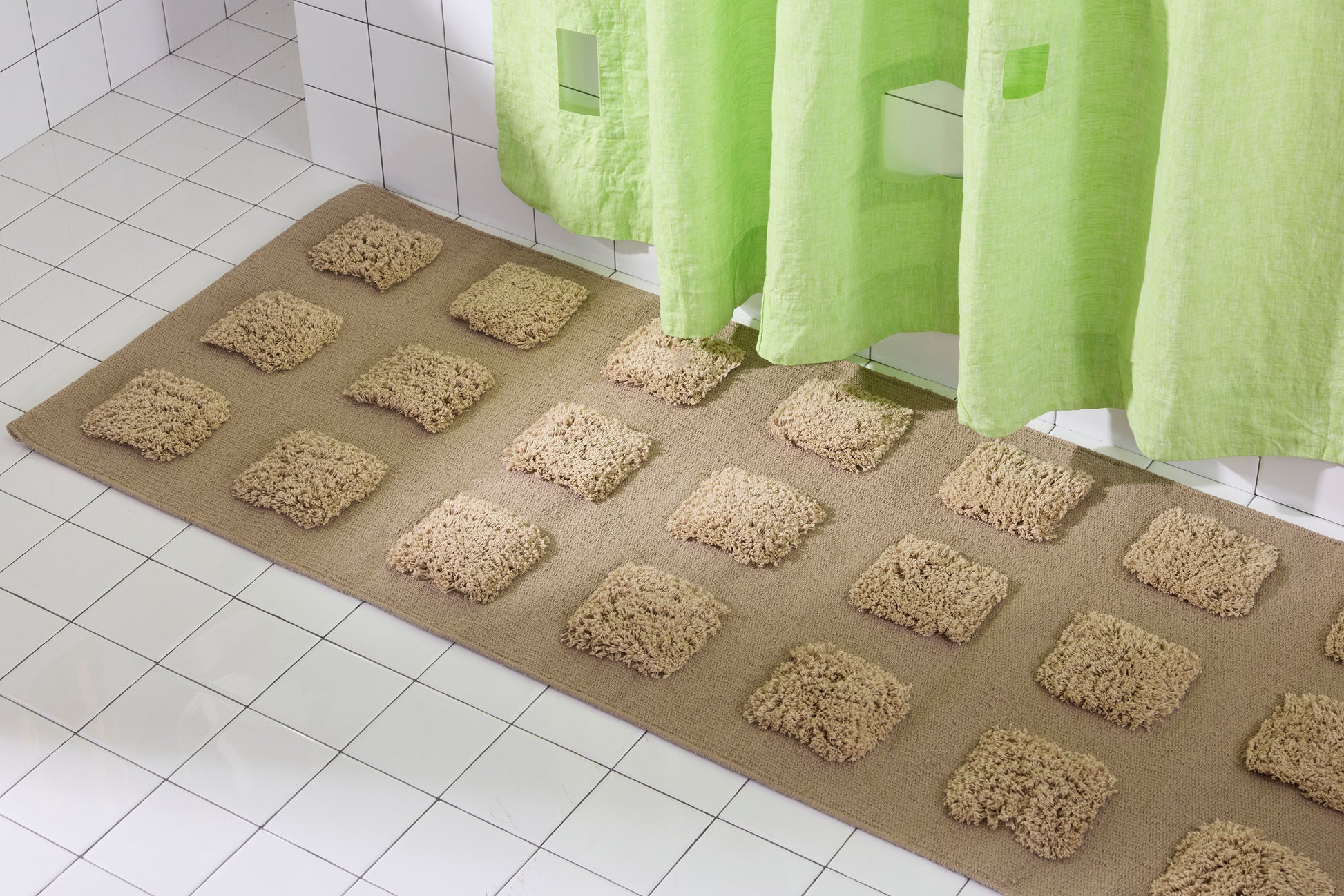 A brown/taupe, natural bath runner with an organic grid of raised tufted squares in warm neutral tones, shown in a bright white tiled bathroom with a lime green shower curtain.