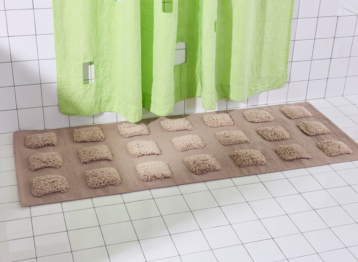A brown bath mat with raised rectangular tufts, shown on a white tiled bathroom floor beneath a lime green shower curtain.