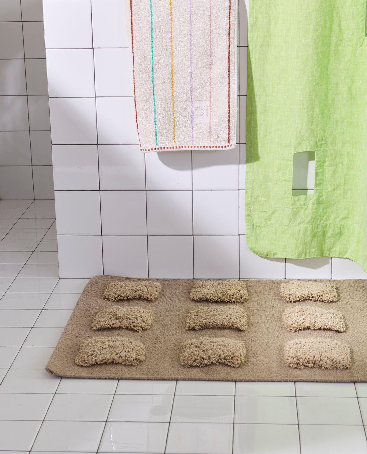 A brown/taupe, natural bath rug featuring rows of raised shaggy tufted squares in varied natural tones, styled in a white tiled bathroom.