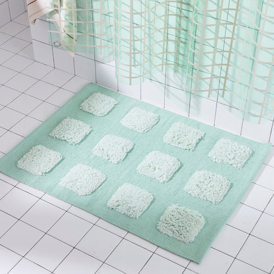 A mint green bath mat with a grid pattern of raised, fluffy square cushions, shown on a white tiled bathroom floor in front of a shower curtain.