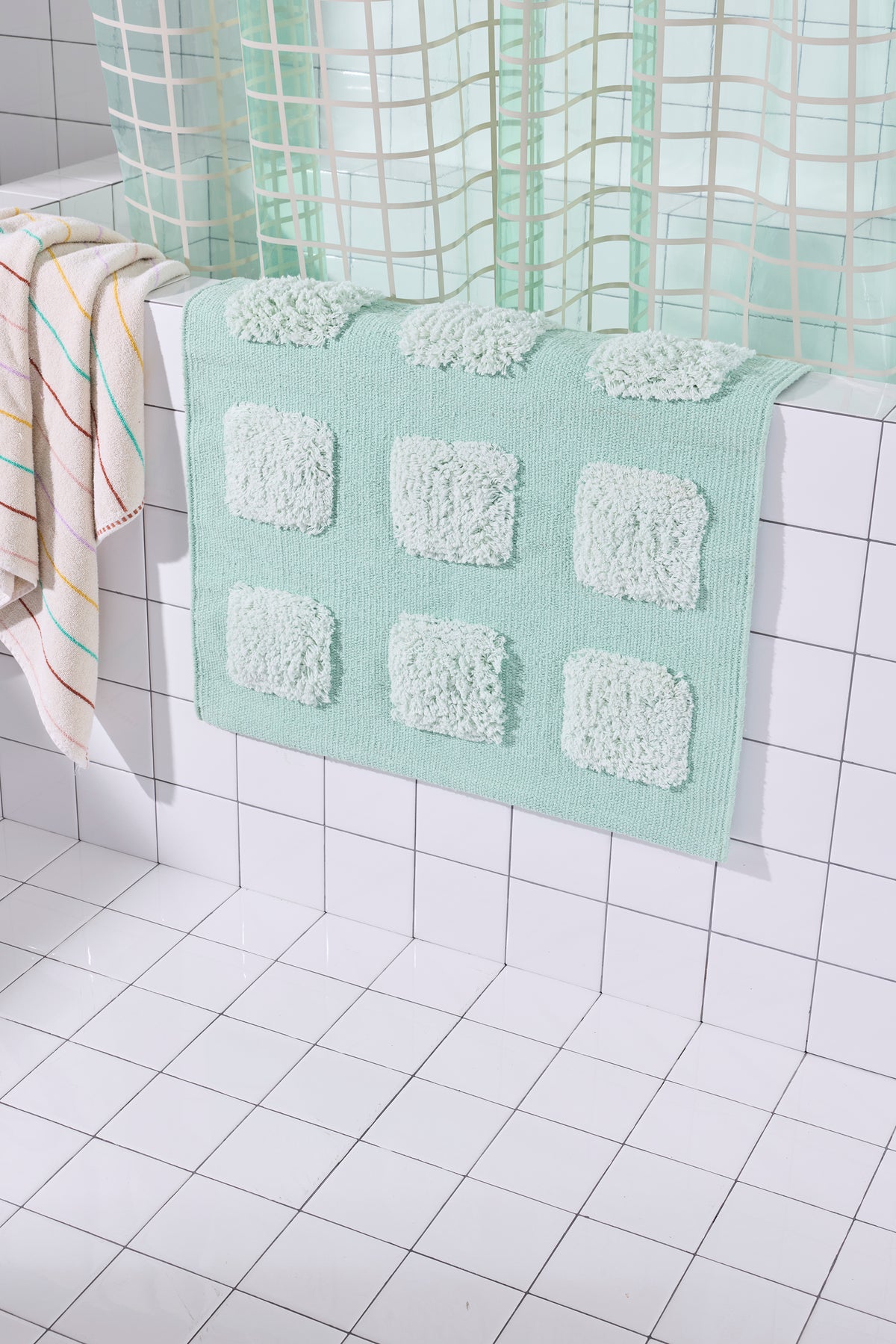 Mint/Teal bath rug with raised tufted squares in natural tones, draped over a white tiled bathroom ledge beneath a teal shower curtain.