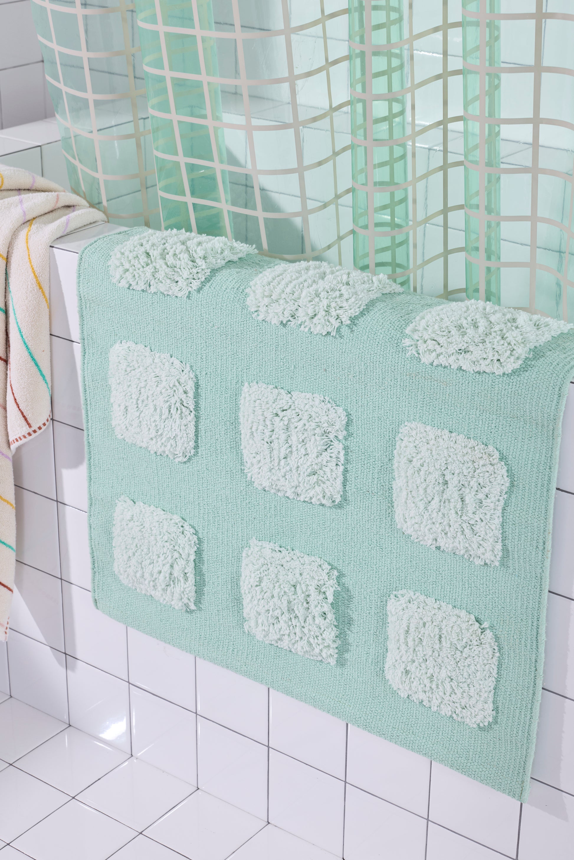 Close up of Mint/Teal bath rug with raised tufted squares in natural tones, draped over a white tiled bathroom ledge beneath a teal shower curtain.