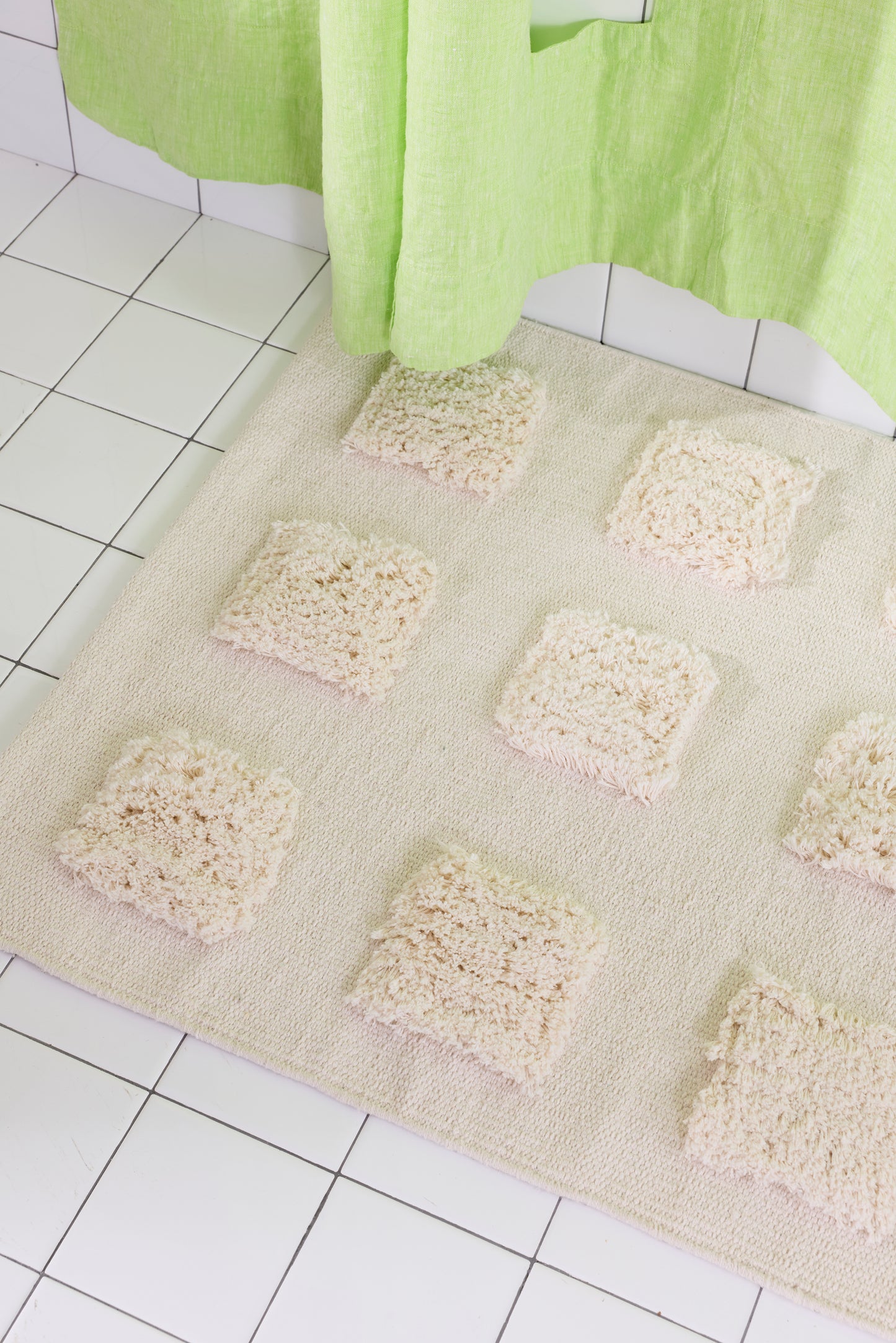 Close-up of the cream, natural bath runner showing its textured woven base and raised plush tufted squares in warm neutral shades beneath a lime green shower curtain.