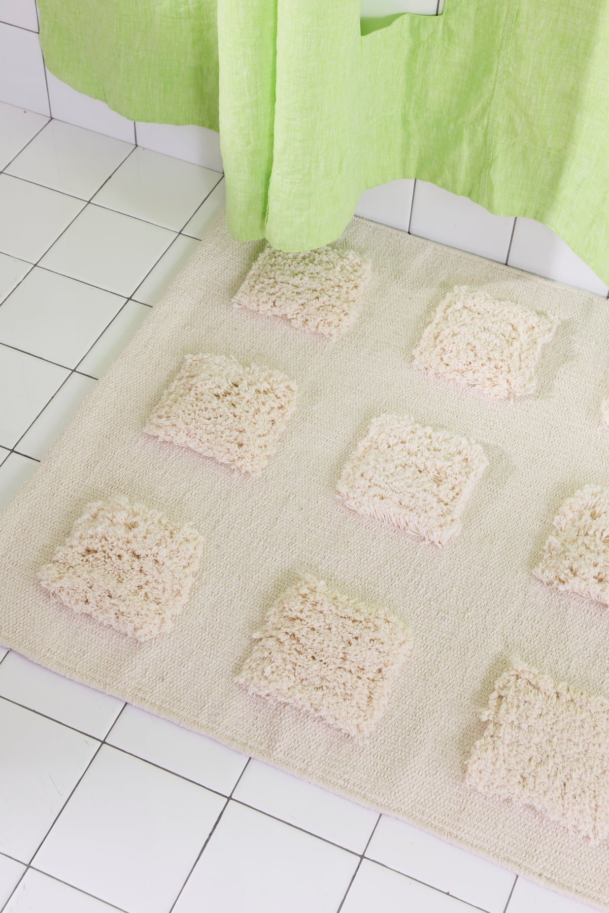 Close-up of the cream, natural bath runner showing its textured woven base and raised plush tufted squares in warm neutral shades beneath a lime green shower curtain.